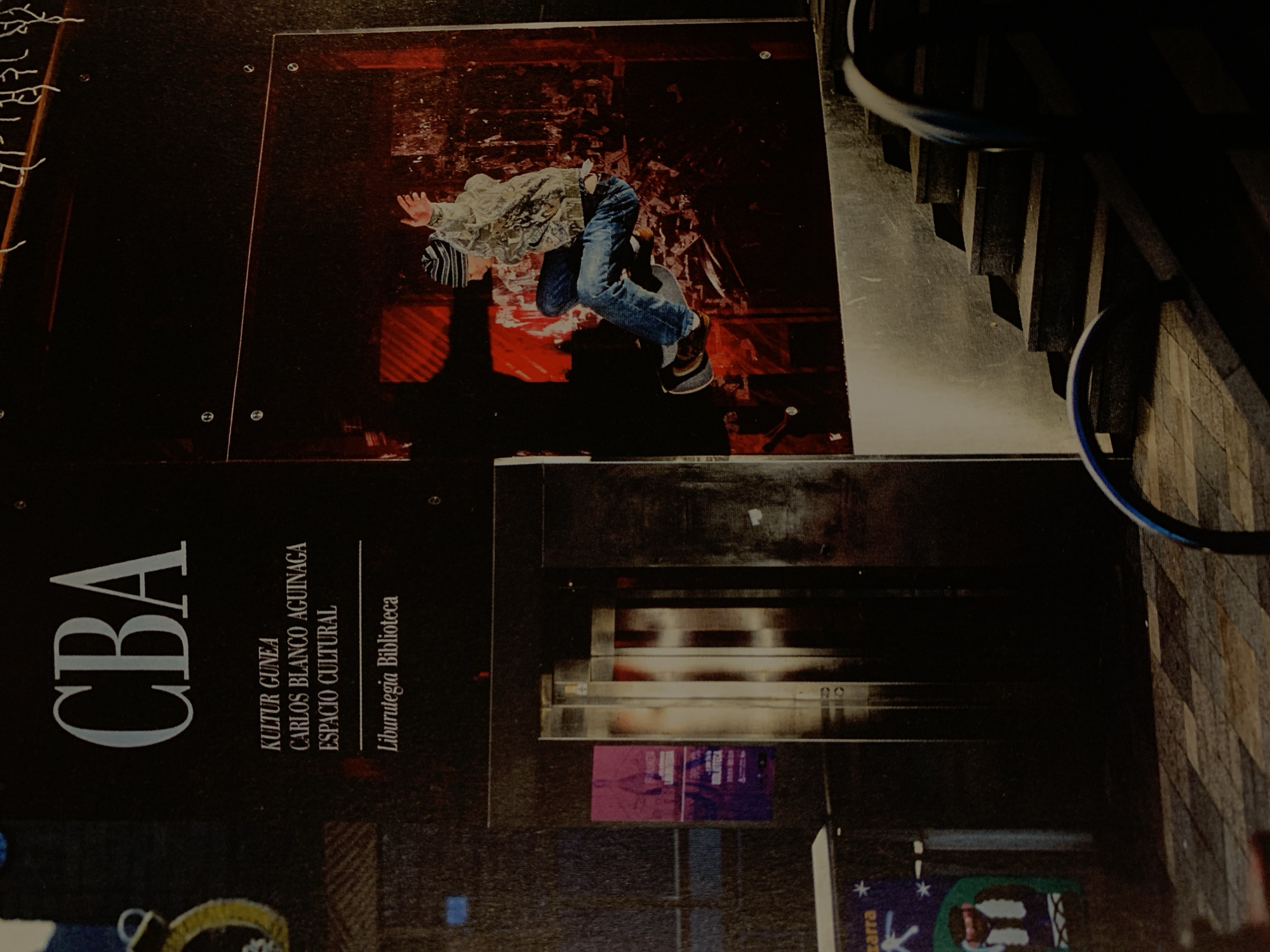 Here's a description of the image:
The image presents an indoor scene that combines elements of art and infrastructure. At the top of the frame, there's an advertisement or display featuring a person in mid-air, possibly performing a jump or acrobatic move. The person is wearing casual clothing, including jeans, a jacket, and a hat. The background of this advertisement is a mix of red and darker tones.
Below this, there's a sign or advertisement for "CBA" (potentially Centro de la Biblioteca Argentina), which includes the text "KULTUR GUNEA", "CARLOS BLANCO AGUINAGA", "ESPACIO CULTURAL", and "Liburutegia Biblioteca".
Adjacent to these elements are parts of an architectural structure, specifically stairs with a curved handrail. The stairwell area appears to be relatively dark, with lighting playing a significant role in defining shapes and textures.