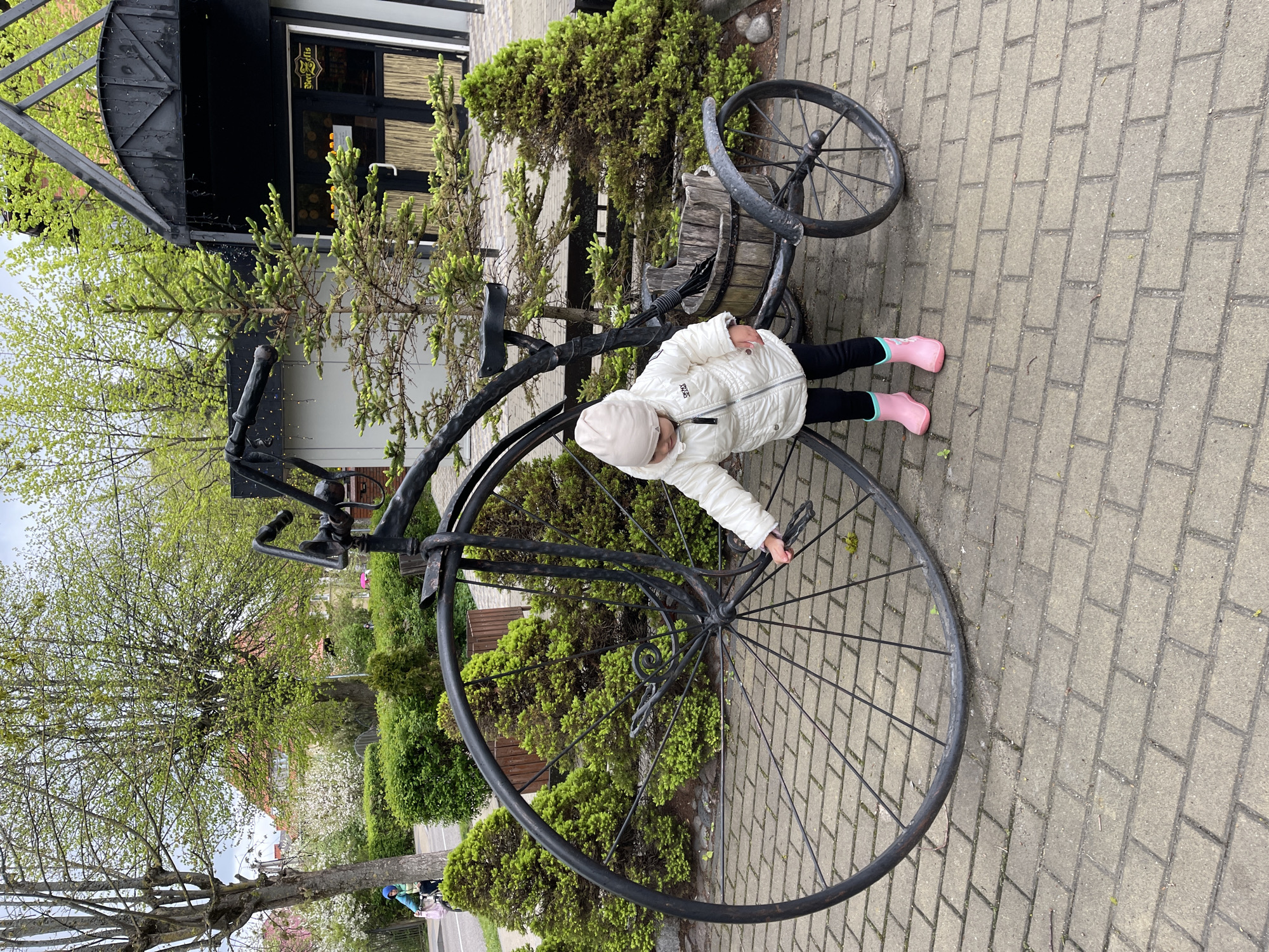 A young child is positioned beside a large, antique-style bicycle on a paved surface, likely a sidewalk. The child wears a white jacket, black pants, a light-colored hat, and pink boots, and is leaning towards the bicycle's large front wheel. The bicycle appears to be a decorative piece or an art installation. The background includes green bushes and trees, suggesting a park or garden setting. A building is visible in the background, with a black awning-like structure and a sign above a window. The location is Zelenogradsk, Russia.