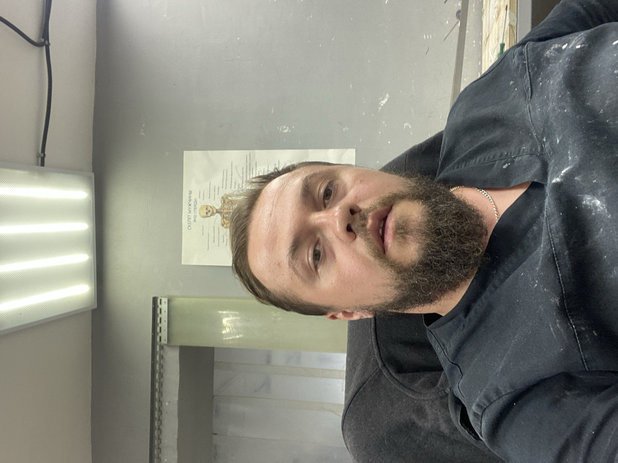 A vertical, medium shot captures a man's face and upper body, his head tilted slightly to the right, suggesting a casual or candid moment. He has short brown hair, a full beard, and is looking directly at the camera with a neutral expression. He wears a dark, collared shirt, possibly a scrub top or work uniform, which is dusted with white specks, hinting at a workshop or studio environment. A thin, silver chain necklace is visible around his neck.
Behind him, a gray wall serves as the backdrop. A medical chart, featuring a diagram of a human skeleton, is pinned to the wall on the left. The text on the chart appears to be in Cyrillic script and reads "СКЕЛЕТ МУЖЧИНЫ ВИД С ПЕРЕДИ" which translates to "MAN'S SKELETON VIEW FROM THE FRONT". To the left of the chart, a long, fluorescent light fixture with a textured diffuser emits a bright, white light. The overall lighting suggests an indoor setting, possibly during the day given the intensity of the light. The presence of the skeleton chart and the white dust on his clothing might indicate he works in a medical, artistic, or perhaps construction-related field. The scene is somewhat cluttered, with a glimpse of shelving units and building materials in the background, further reinforcing the idea of a workspace.