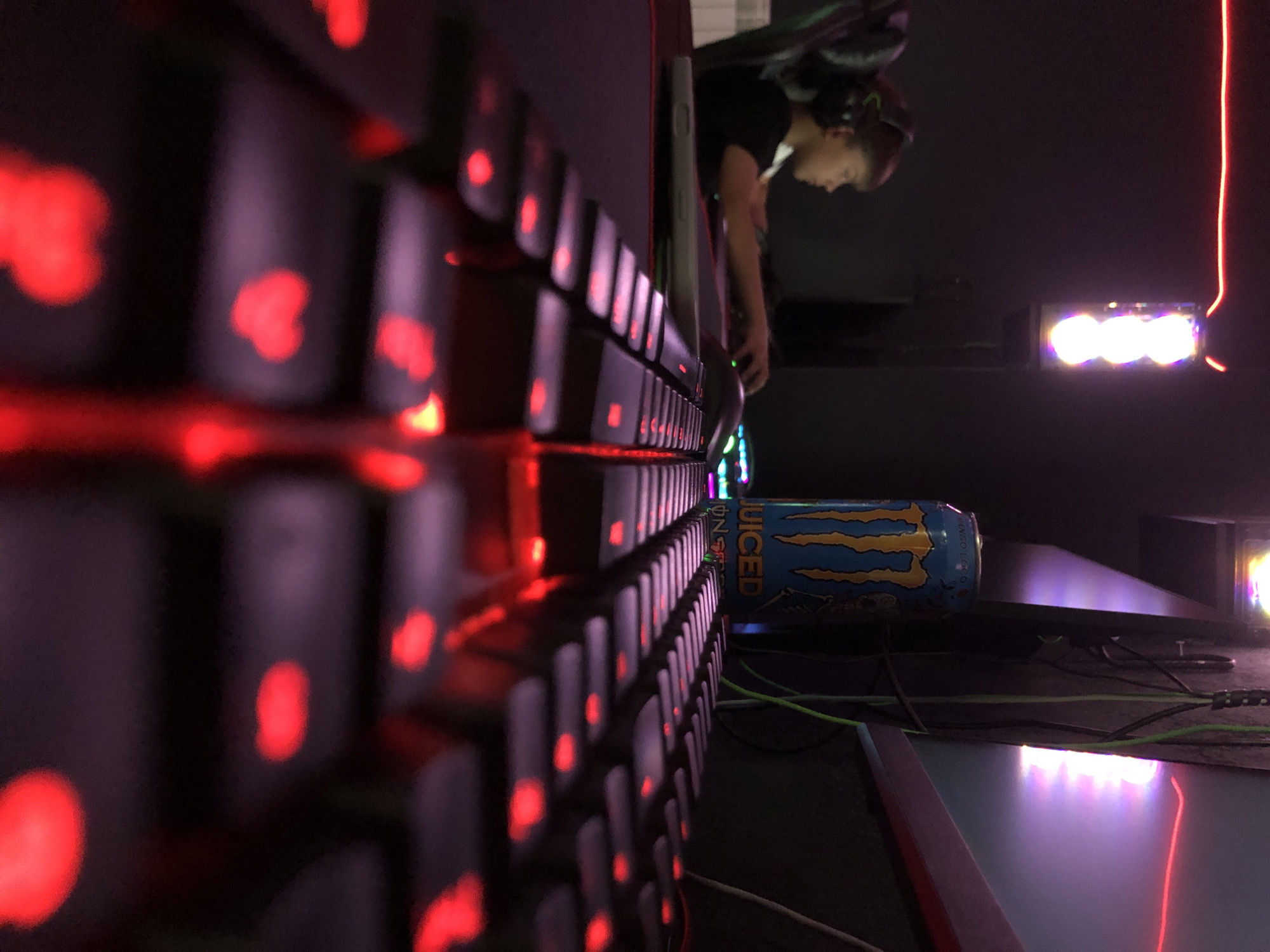 The scene, set in Beryozovka, Russia, depicts a dimly lit indoor gaming environment. In the foreground, a black mechanical keyboard with prominent red LED backlighting is angled upwards, occupying the lower half of the frame. The red backlighting illuminates several keycaps, some in sharp focus, others blurred due to the shallow depth of field. A keycap featuring a heart shape is visible on the lower left section of the keyboard.
Behind the keyboard, on a dark desk surface that appears to be covered by a mouse pad with a red border, a blue Monster Energy "Juiced Mango Loco" can is prominently displayed. To the left of the can, a white rectangular object, possibly a smartphone or tablet, rests on the desk. A black gaming mouse with subtle RGB lighting is partially visible to the right, beyond the keyboard's focus.
In the midground, a young male individual is seated in a dark gaming chair, wearing black over-ear headphones and a black t-shirt. He is positioned facing right, with his right hand operating a computer mouse, indicating active computer use or gaming.
The background features various artificial light sources: a vertical fixture on the left illuminates with three distinct, bright, circular white-to-purple lights. A thin, horizontal red LED strip light runs across the upper left corner of the room. The area to the right, behind the monitor (which is partially obscured), emits a soft purple-pink glow. Green electrical cables are visible running vertically along a dark structure behind the monitor and the energy drink can. The surrounding room walls appear dark and indistinct.