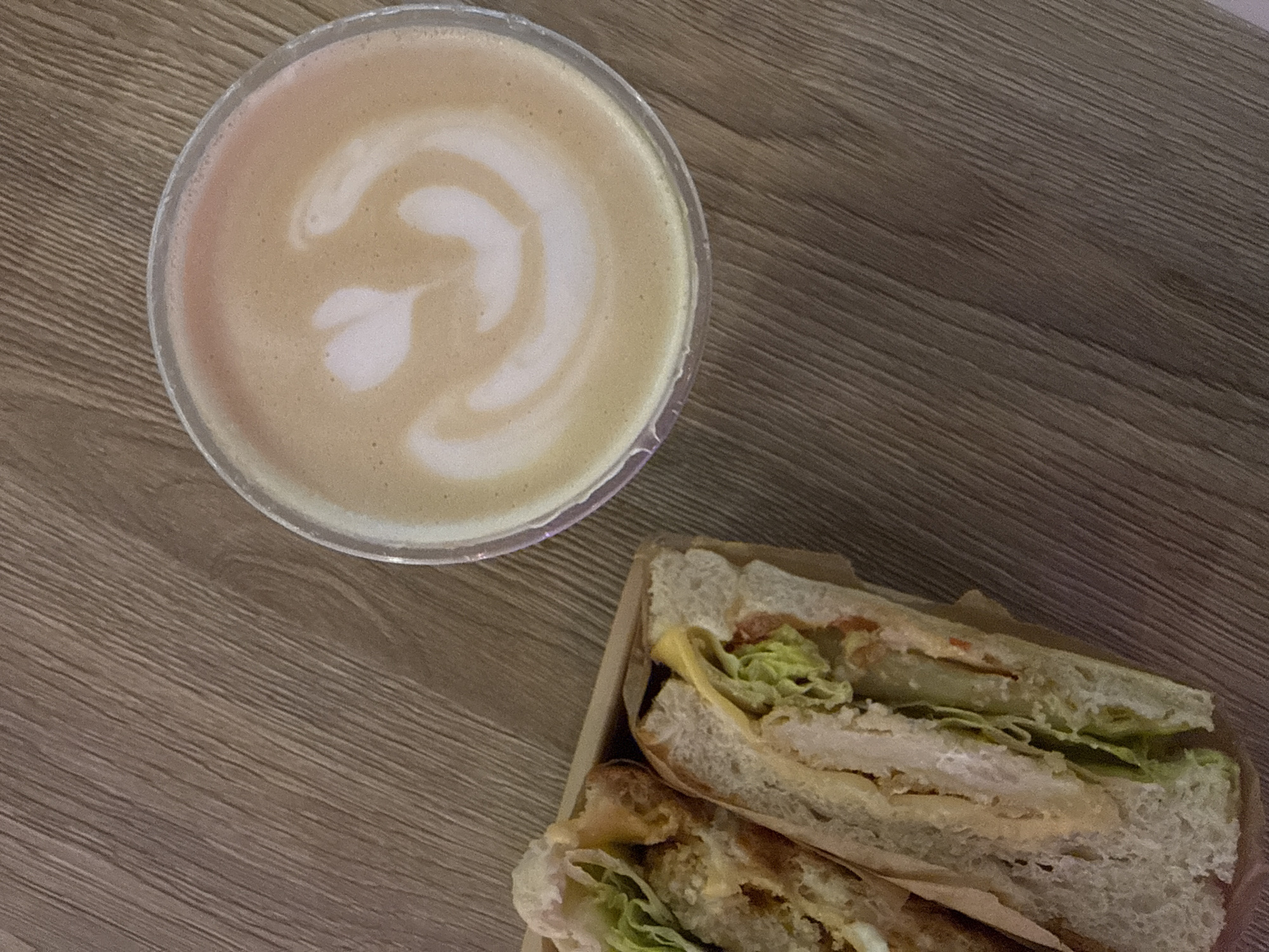 This image captures an overhead view of a coffee drink and a sandwich resting on a wooden table.
The main subject on the left is a clear plastic cup filled with a latte or similar milk-based coffee. The surface of the coffee features delicate white latte art, forming a distinct heart shape within a swirl pattern against the light brown coffee foam.
To the right, a sandwich, cut in half, is presented in a light brown cardboard tray or box. The sandwich appears to be a sub or baguette-style roll, generously filled. Visible ingredients include fresh green lettuce, slices of red tomato, yellow cheese, and what looks like pieces of cooked chicken or meat, possibly breaded. A creamy sauce is also discernible among the fillings. The bread is white and appears soft.
The items are placed on a wooden surface, likely a table, which displays a distinct light brown color with a prominent diagonal wood grain pattern. The lighting is soft and even, suggesting an indoor setting.
There are no people visible, and therefore no discernible emotions. The time of day cannot be determined from the image itself, though coffee and sandwiches are typically consumed during breakfast, brunch, or lunch. No visible text is present on any of the objects. While the image provides no internal cues to location, the provided context indicates the location is Moscow, Russia.