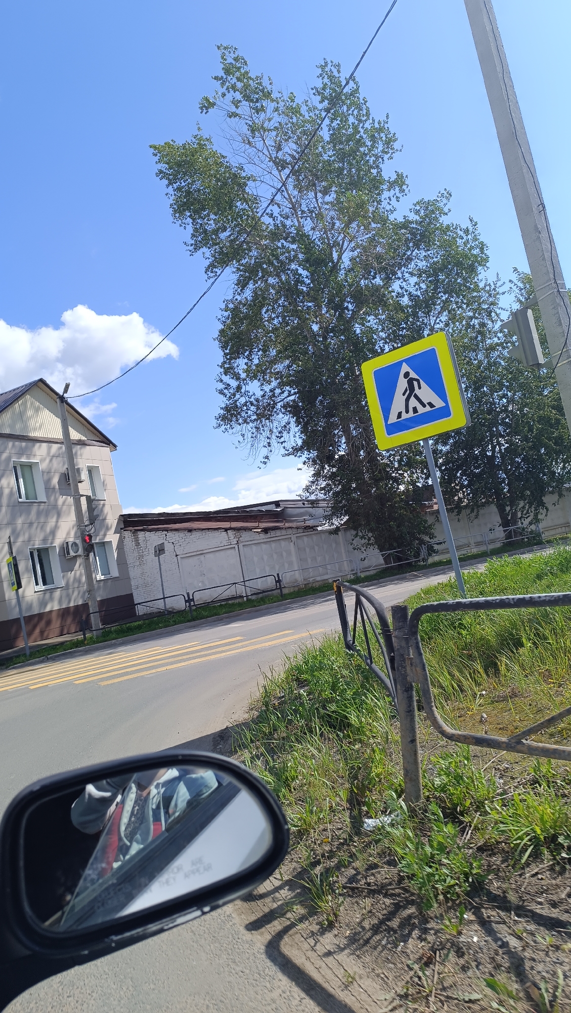 A view from a vehicle in Chaikovsky, Russia, shows a residential building with a traffic light on the left, a white wall with a fence, a tree, and a pedestrian crossing sign. The pedestrian crossing sign is a blue square with a yellow border and a black silhouette of a person walking. The traffic light is red. The residential building appears to be made of concrete and has several windows. The white wall is approximately 10 feet tall and stretches across the background. A large tree with green leaves is positioned behind the building and wall. The sky is blue with some white clouds. The road in front of the building is a pedestrian crossing with yellow stripes. The image is taken during the day, and the sun is shining. The reflection in the vehicle's side mirror shows a person wearing a light-colored hoodie.