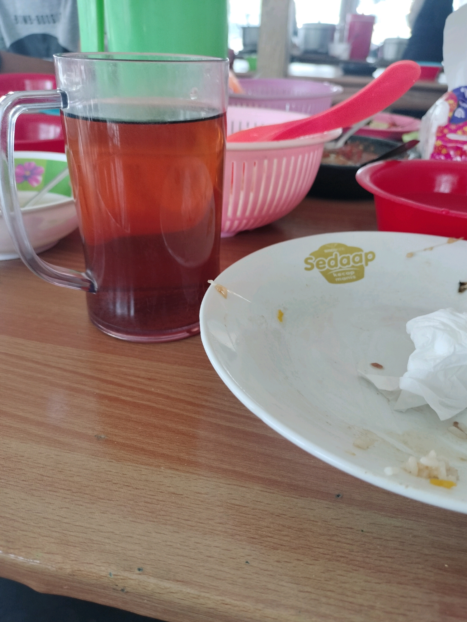 A close-up, eye-level, indoor, slightly right-of-center shot shows a glass mug filled with a dark amber liquid, likely tea, resting on a wooden table. The mug has a clear handle and the liquid occupies approximately two-thirds of its volume. To the right of the mug is a white plate with remnants of food, including rice and a crumpled white napkin. The plate has a yellow logo with the text "Sedaap kecap manis". In the background, out of focus, are various colorful objects including pink and green plastic containers and a red bowl. Part of a person's gray t-shirt is visible in the upper left corner. The overall scene suggests a casual dining setting, possibly at a food stall or restaurant. The location is specified as Gambah Luar, Indonesia.