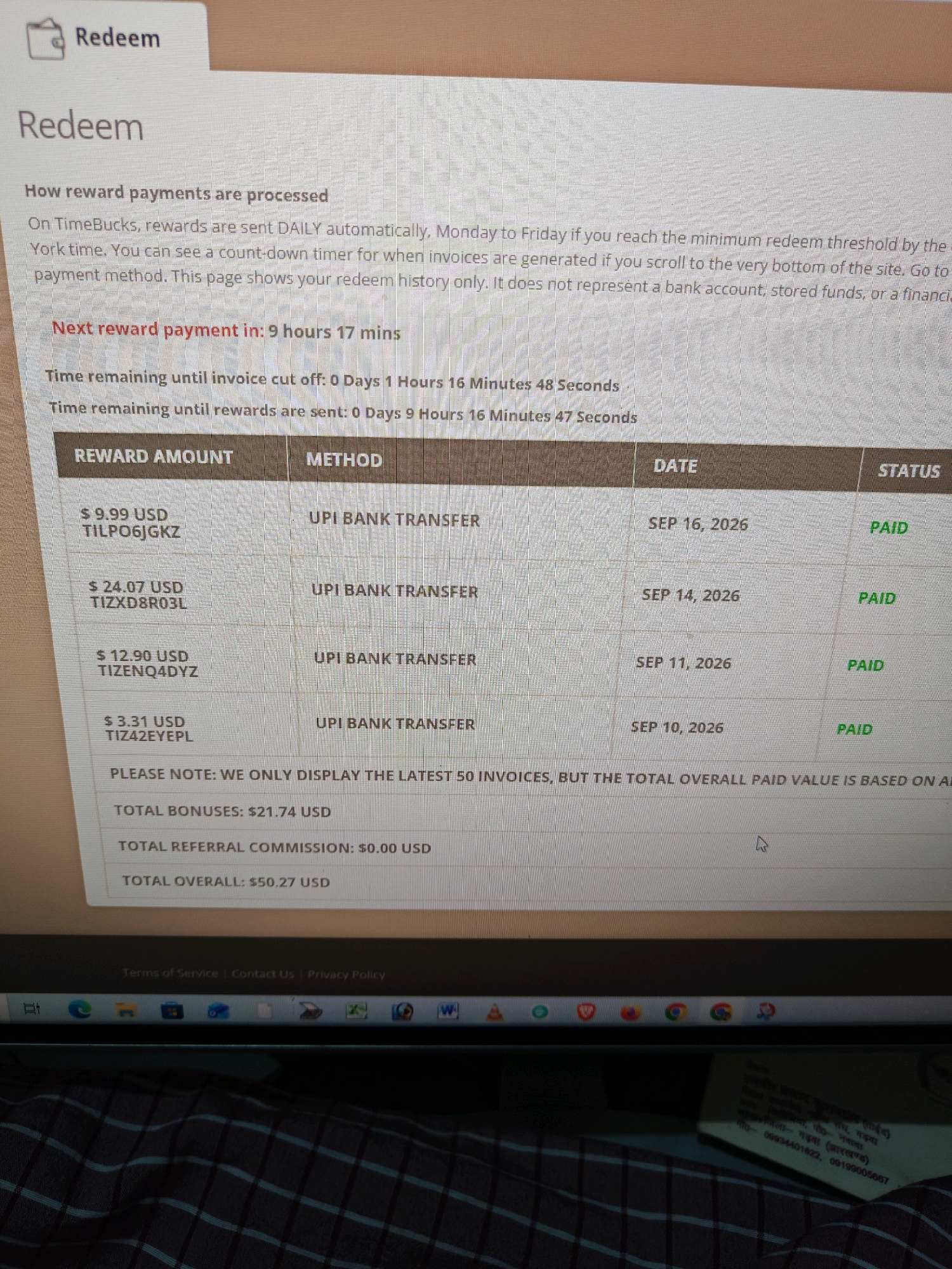 The image displays a computer screen showing a "Redeem" page from a website called "TimeBucks." The page details how reward payments are processed, showing a countdown timer for the next reward payment and the time remaining until invoice cut-off and rewards are sent. Below this, a table lists past reward transactions, including reward amount, method (UPI BANK TRANSFER), date, and status (all "PAID"). The total bonuses, total referral commission, and total overall paid value are also summarized. The bottom of the screen shows the taskbar of a Windows operating system, with various application icons and the system clock. Part of a folded piece of paper with text in Hindi is visible below the screen, partially obscuring the lower part of the computer's bezel. The context provided in the prompt about the location in Garhwa, India, is not discernible from the image itself, as the image only shows a digital interface and a small portion of an unrelated physical object.