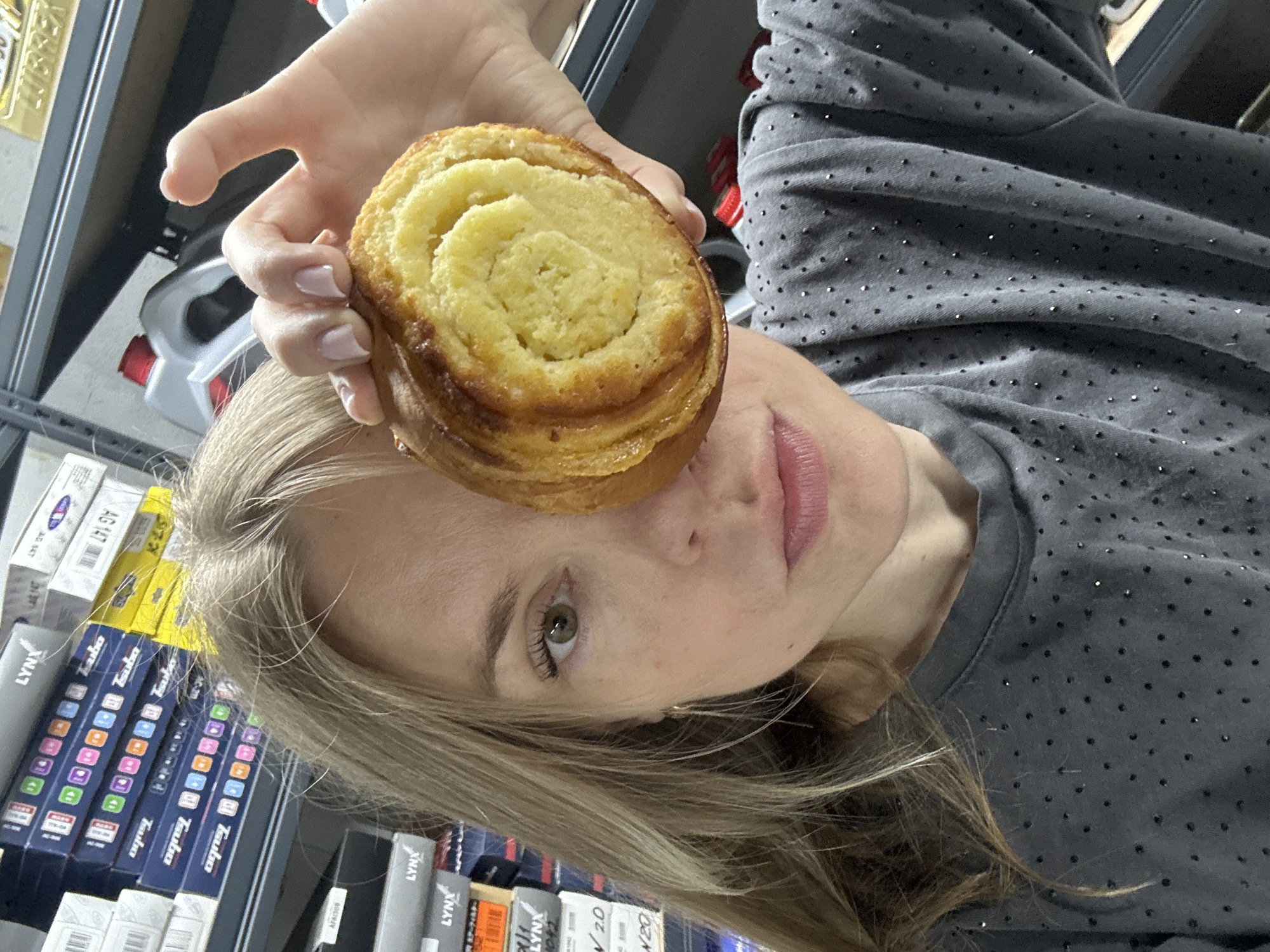 A woman is holding a round pastry in front of her right eye, obscuring it from view. She has blonde hair, fair skin, and is wearing a grey t-shirt adorned with small, dark embellishments. The background appears to be a storage area with shelves stacked with boxes. The image does not provide any specific information about the location in Blagoveshchensk, Russia.