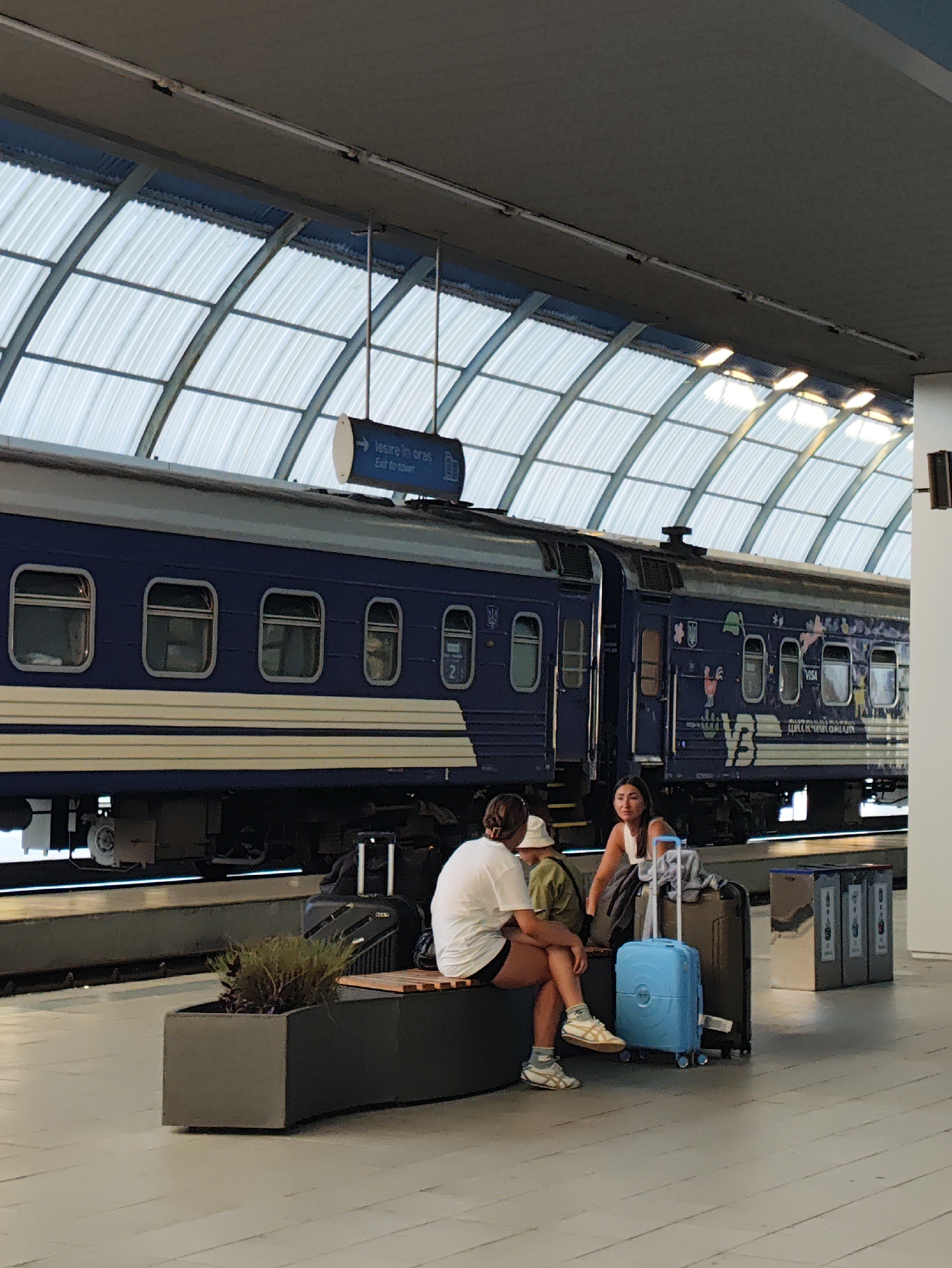 The image depicts a train station platform in Chișinău, Moldova. A blue train with white stripes is parked at the platform. Several people are seated on a bench, with luggage around them, including a bright blue suitcase and a dark brown suitcase. Above the train, a sign points to "Ieșire în oraș" (Exit to town). The platform roof is made of a translucent, curved material, with fluorescent lights visible. A planter box with greenery is situated in front of the bench. The floor is paved with light-colored tiles.