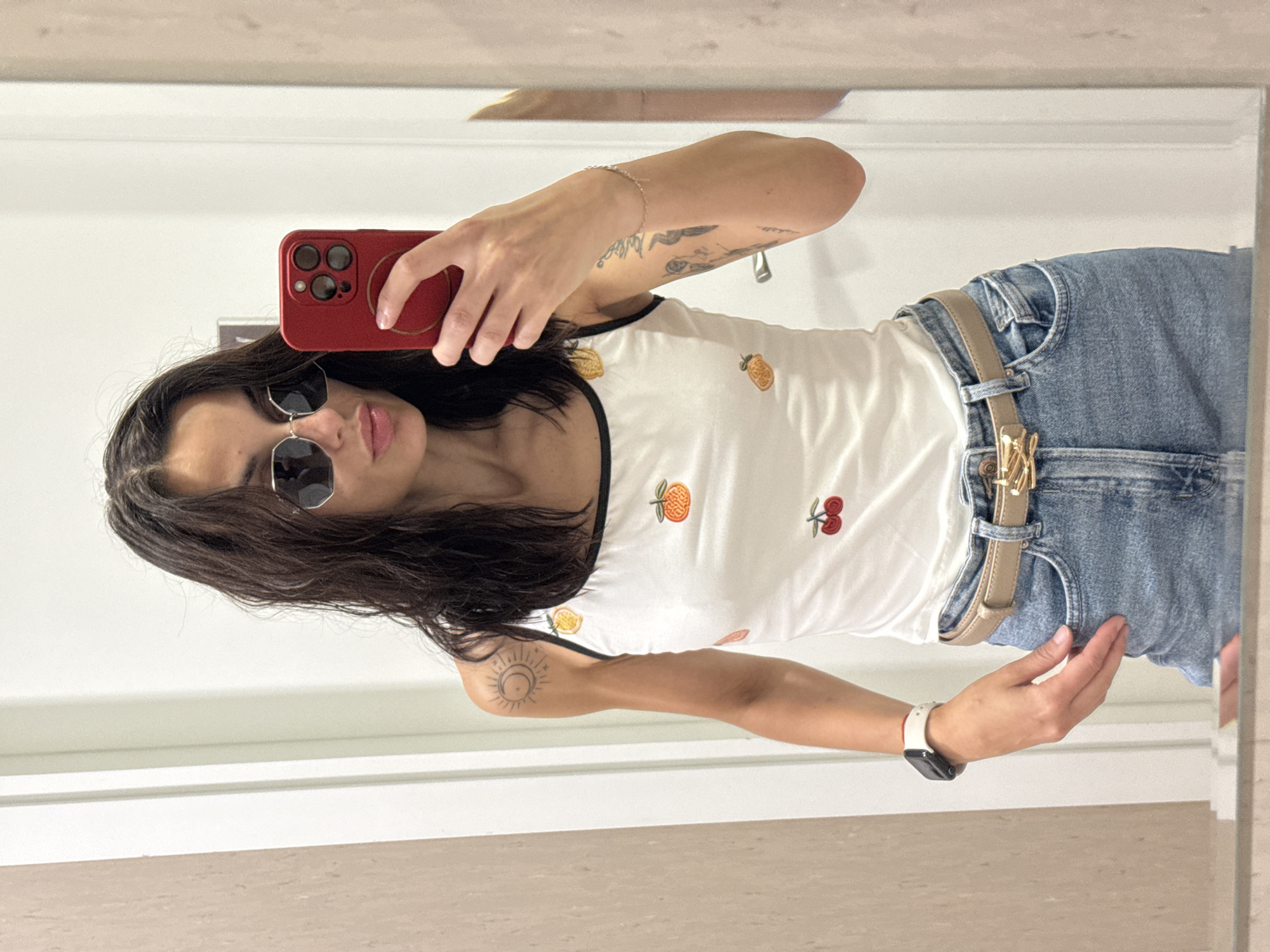 A female individual is captured in a mirror selfie in an indoor setting in Pleven, Bulgaria. She has dark, wavy hair and is wearing round, dark-lensed sunglasses, with her lips slightly parted. Her attire consists of a white sleeveless top embellished with embroidered fruit designs (oranges and cherries) and light blue denim jeans. A light brown belt with a distinctive gold, abstract-shaped buckle is worn around her waist. On her left wrist, she wears a white smartwatch. Her right wrist displays a thin, light-colored bracelet, and she holds a red smartphone, equipped with a circular grip accessory, to capture the image. Tattoos are visible on both arms: a sun and crescent moon design on her left upper arm/shoulder, and an indistinct tattoo on her right forearm. The reflected background reveals a white wall with a vertical white door frame on the right side. A light brown or beige textured wall surface is visible on the left side of the reflection. The scene is uniformly lit.