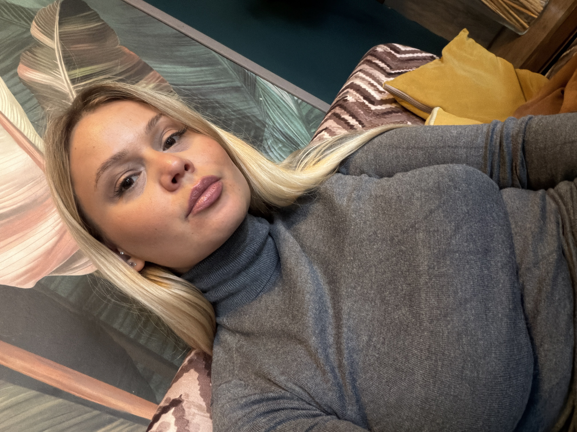 A blonde woman with light skin is the subject of this selfie. She has brown eyes, full pink lips, and wears a gray turtleneck sweater. Her hair is parted to one side and falls around her face. She appears to be lying down, as indicated by the angle and her relaxed posture.
The background includes a wall with a stylized feather pattern in muted pink and green tones. Part of a striped cushion in brown and white, along with a yellow pillow, are visible to the upper right.
The image does not contain any information to indicate that it was taken in Sarapul, Russia. The location is unknown.