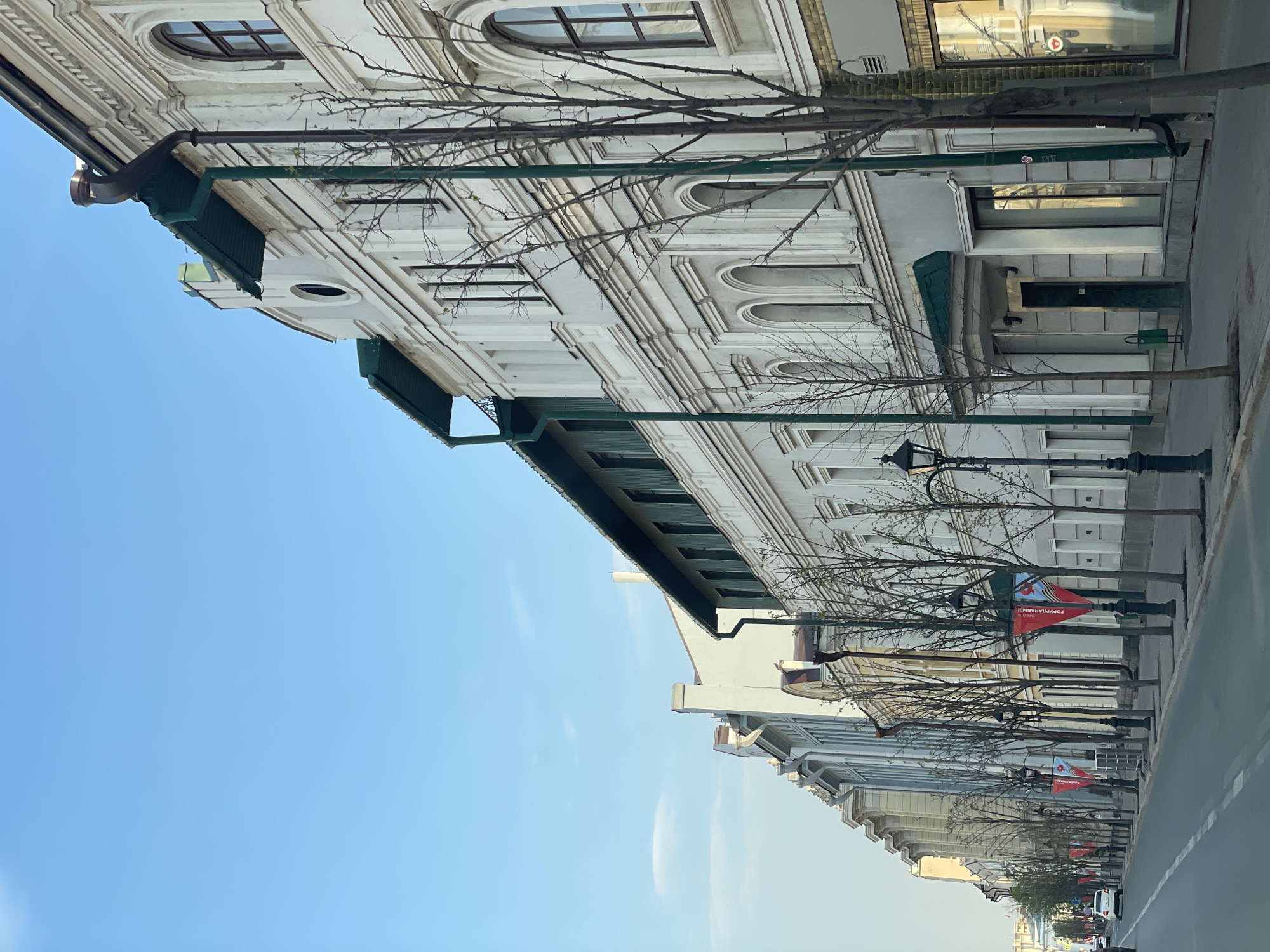 The image depicts a street in Kazan, Russia, lined with ornate, multi-story buildings with classical architectural elements, including arched windows, decorative moldings, and green awnings. Tall, bare trees stand between the buildings and the sidewalk, some displaying red banners. Street lamps are positioned along the sidewalk. The sky is blue. The scene appears to be taken from inside a vehicle.