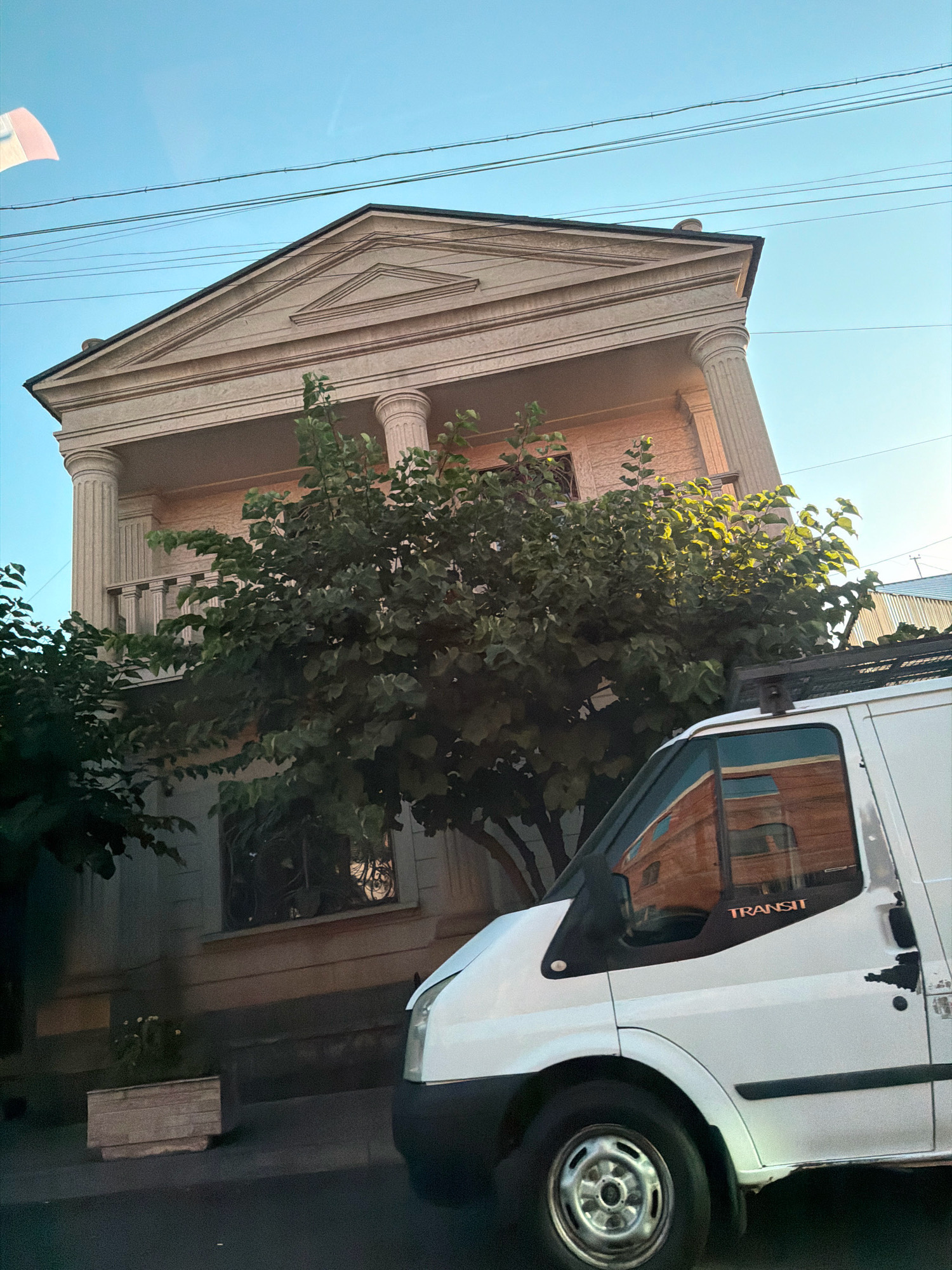 The image captures a white Ford Transit van parked on a street in Yerevan, Armenia, with a classical-style building behind it. The van is angled, showing its front passenger side, including the wheel, fender, door, and side mirror. The building features columns and a triangular pediment, suggesting a grand architectural style. A large tree with dense foliage partially obscures the building and the van. Electrical wires are visible against a clear blue sky. The overall scene depicts an urban environment with a juxtaposition of modern transportation and traditional architecture.
