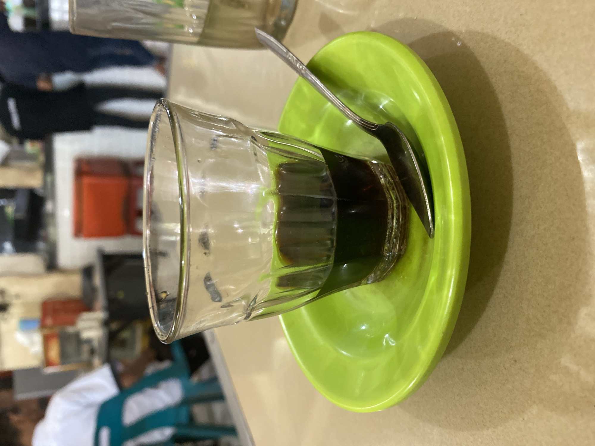 A close-up shot focuses on a small glass of dark liquid, presumably coffee, resting on a bright green saucer. A silver spoon lies on the saucer, partially submerged in the liquid. The glass has a ribbed texture, and the liquid fills the bottom third of the glass. The scene is set on a tan, textured surface that appears to be a tabletop. In the blurred background, two individuals are visible. One person, wearing a white shirt and sitting on a teal chair, is facing away from the camera. Another person, wearing a dark jacket, stands in the background. Behind them, there are hints of a shop or cafe interior, with shelves and colorful containers visible. The image was taken in a cafe in Lhokseumawe, Indonesia.