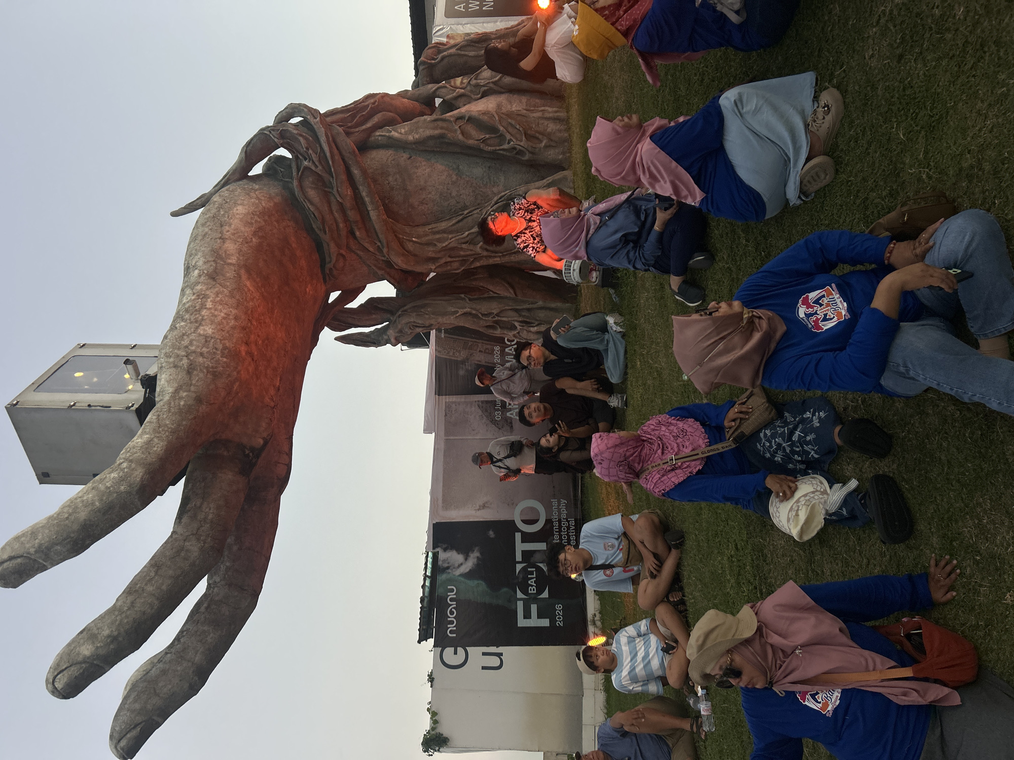A group of people are sitting on a grassy lawn, watching a performance or presentation. In the background, a large, abstract sculpture of a hand and tree-like structure is illuminated by red light. Behind the sculpture is a large white banner with text and images. The sky is clear and pale, suggesting it is either early morning or late afternoon. The people are dressed casually, and some are wearing headscarves. The overall atmosphere appears relaxed and communal. The scene is not identifiable as being in Kediri, Indonesia.