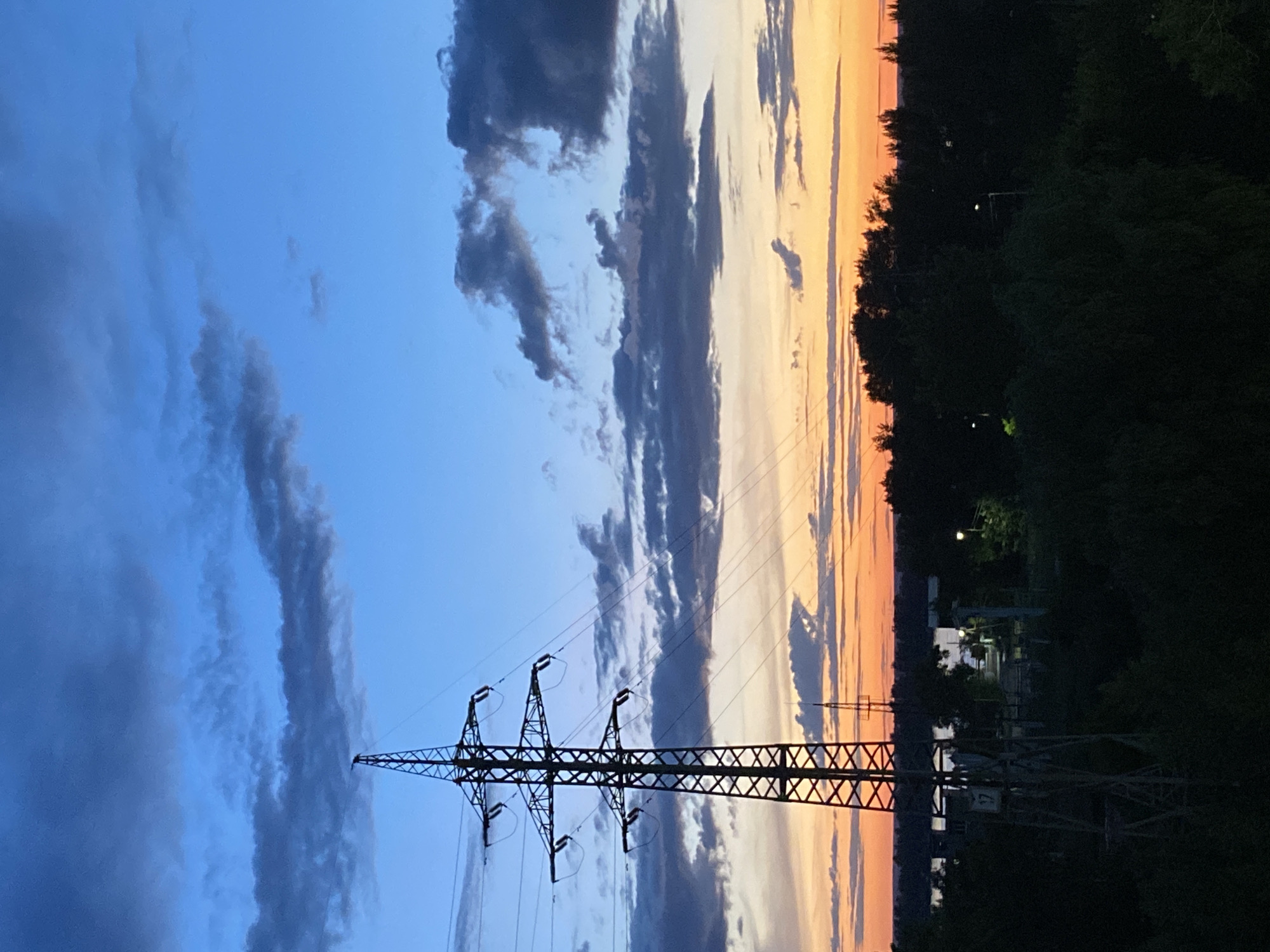 The image depicts a sunset scene in Novokuybyshevsk, Russia. A tall, lattice-like power transmission tower dominates the left side of the frame, with its silhouette stark against the colorful sky. Several power lines extend from the tower and disappear out of frame. The sky transitions from a deep blue at the top, with scattered dark clouds, to hues of orange and yellow near the horizon, indicating the setting sun. Below the sky, a dark silhouette of trees and foliage spans the lower portion of the image. In the middle ground, a body of water can be seen, with faint lights reflecting on its surface. Further in the distance, more trees and structures are visible, also silhouetted against the sunset. The overall impression is one of a serene, industrial landscape at dusk.