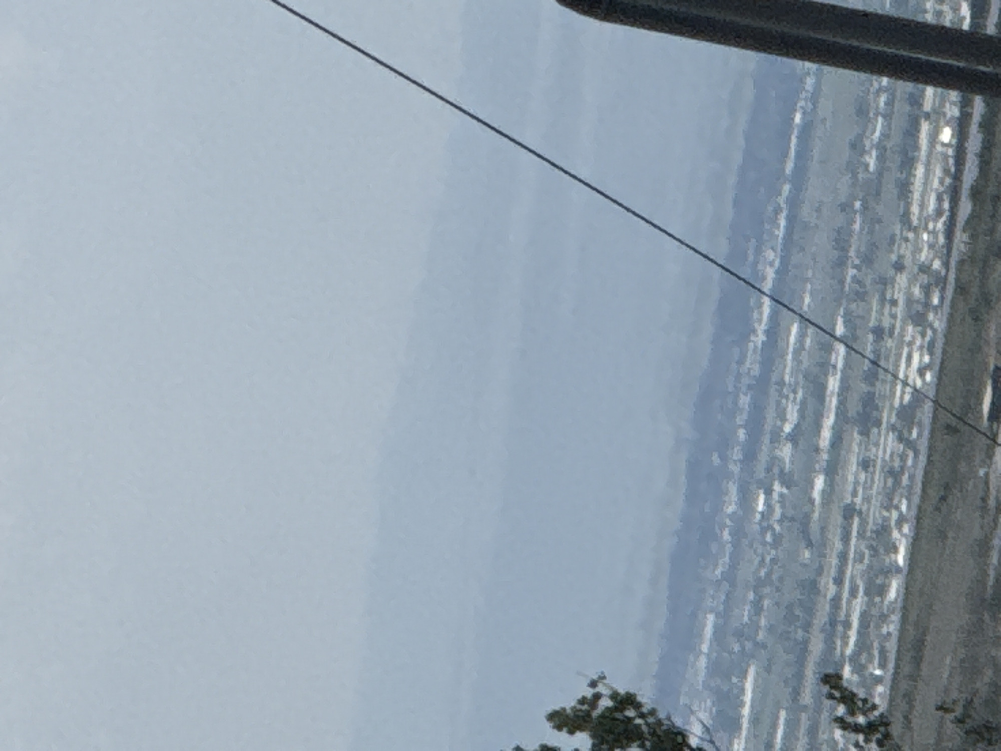 The image displays a panoramic view of a distant urban or suburban landscape under an overcast sky, rotated 90 degrees counter-clockwise from its likely original orientation. When viewed upright, the upper portion is dominated by a light blue-grey, hazy sky.
A thin, dark diagonal line, appearing to be a wire or cable, stretches across the sky from the top left towards the middle right. In the top right corner, a thick, dark, horizontal bar, possibly part of a pole or a structural element, cuts into the frame.
Below the sky, extending across the middle ground, is a sprawling distant landmass. This area consists of indistinct, blocky shapes in muted grey and blue tones, suggesting a city or developed region with buildings and roads. Some small, bright spots are visible within this distant cityscape, possibly reflections or distant lights. The overall impression is one of a vast, developed area viewed from a high vantage point, with the distance and atmospheric haze blurring specific details.
In the bottom right corner, dark green, blurry shapes resembling foliage or treetops are visible, suggesting nearby vegetation. The lighting is soft and diffuse, characteristic of an overcast day. There are no people, explicit activities, or visible text in the image.