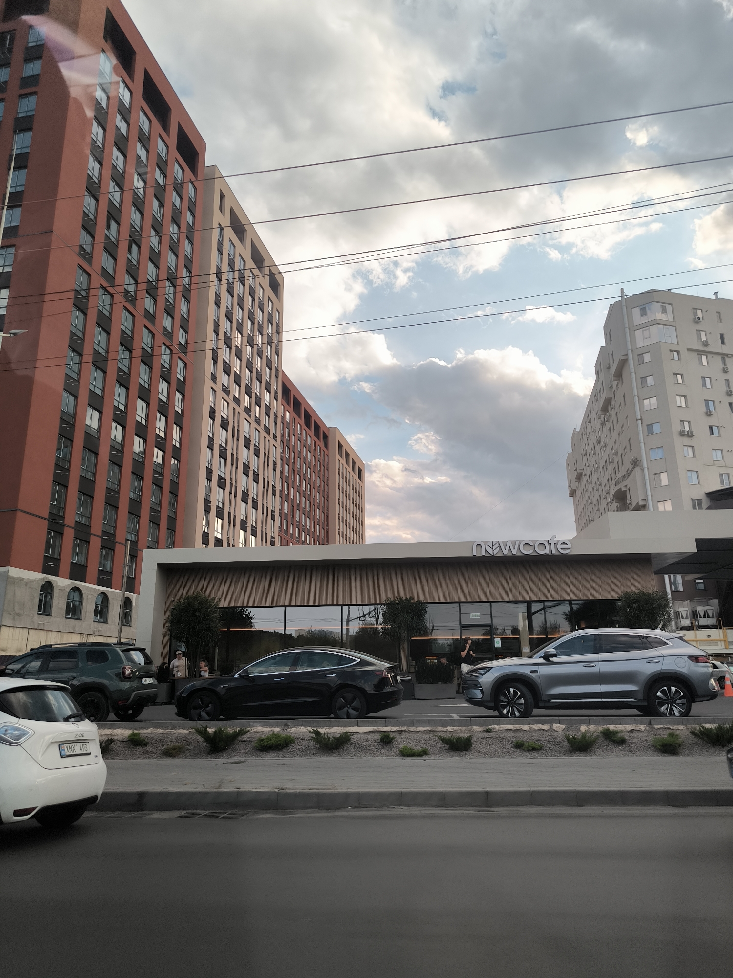 The image depicts the intersection of Ștefan cel Mare și Sfânt Boulevard and Pushkin Street in Chișinău, Moldova. In the foreground, on the right side of the frame, a silver SUV is parked on the street. To the left of the SUV, a black sedan is parked. Further to the left, a white electric vehicle is partially visible. Behind the vehicles, a modern building with a prominent "nwcafe" sign is situated. This building features a glass facade and a wooden-paneled exterior. Two multi-story residential buildings are visible in the background. The sky is partly cloudy, with scattered clouds against a light blue backdrop. Overhead, power lines crisscross the sky.