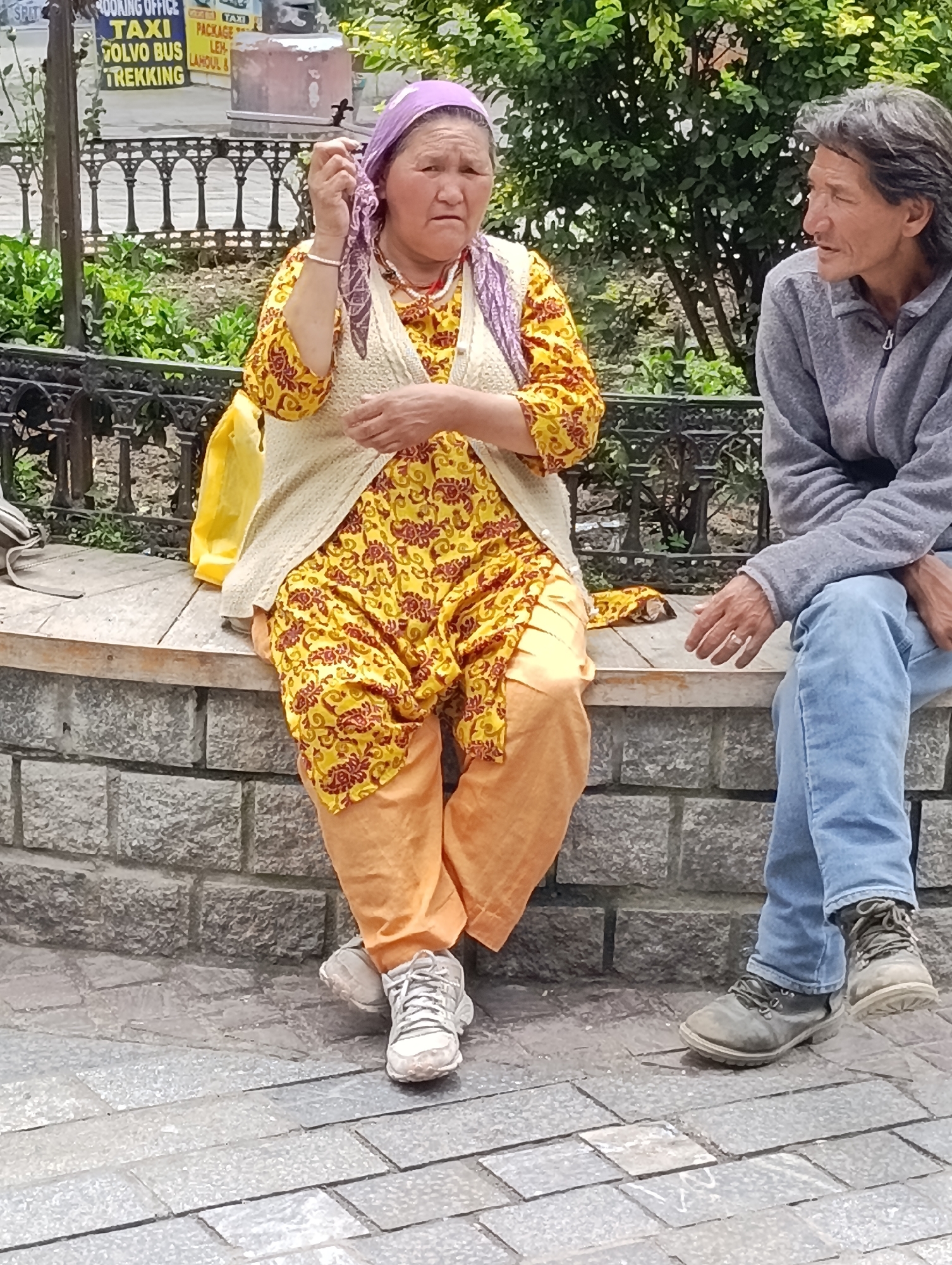 In Manali, India, two individuals are seated on a low stone wall. The person on the left, an elderly woman wearing a purple headscarf, a yellow and red patterned garment, and an off-white vest, is holding a piece of fabric to her face. She is also adorned with necklaces and bracelets. To her right, a person with long, dark hair, dressed in a grey zip-up jacket and blue jeans, sits with their legs crossed. The background reveals a park-like setting with green foliage and trees, and a black metal fence. In the distance, a sign indicates taxi, Volvo bus, and trekking services. The ground is paved with stone tiles.