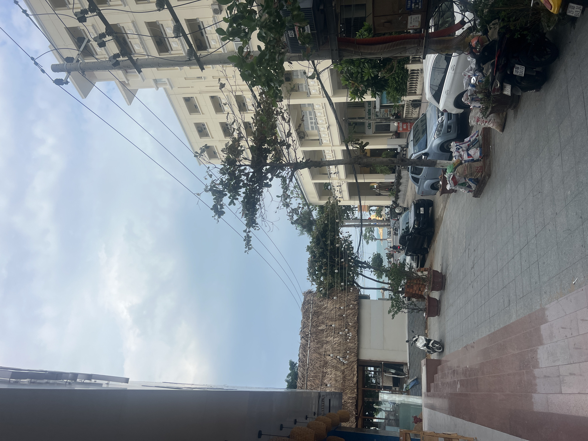 The image depicts a street scene in an urban area, potentially a tourist destination, in an unknown city. On the right side of the image is a sidewalk paved with rectangular tiles, lined with vehicles, including cars and motorbikes. Some parked vehicles appear to be covered with tarps and miscellaneous items. Adjacent to the street, there is a multi-story building with numerous windows and balconies. Power lines crisscross the sky above the street. In the foreground, a structure with a thatched roof element is visible, possibly part of a restaurant or commercial establishment. The sky is overcast with a light blue hue. The image is rotated, with the horizon appearing vertical along the right side.