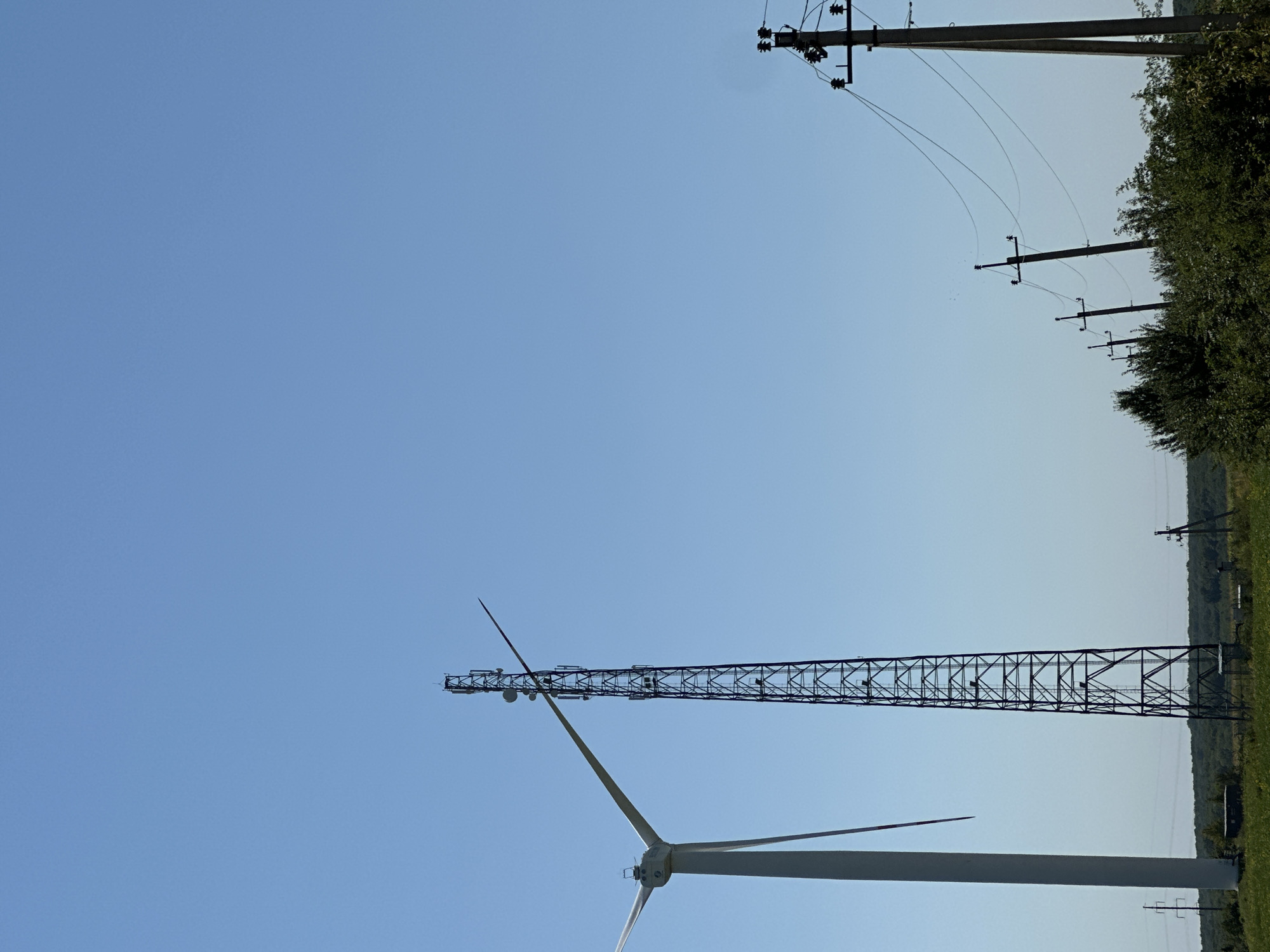 In Korchivka, Ukraine, on a clear, sunny day, a tall, lattice metal tower with multiple antennas stands against a bright blue sky. To the left of the tower, a portion of a white wind turbine with a single blade extended is visible. In the foreground and stretching across the right side of the image, a line of utility poles with electrical wires is present, partially obscured by green foliage. The landscape appears rural, with a line of trees forming a distant horizon. There are no people or animals visible in the scene.