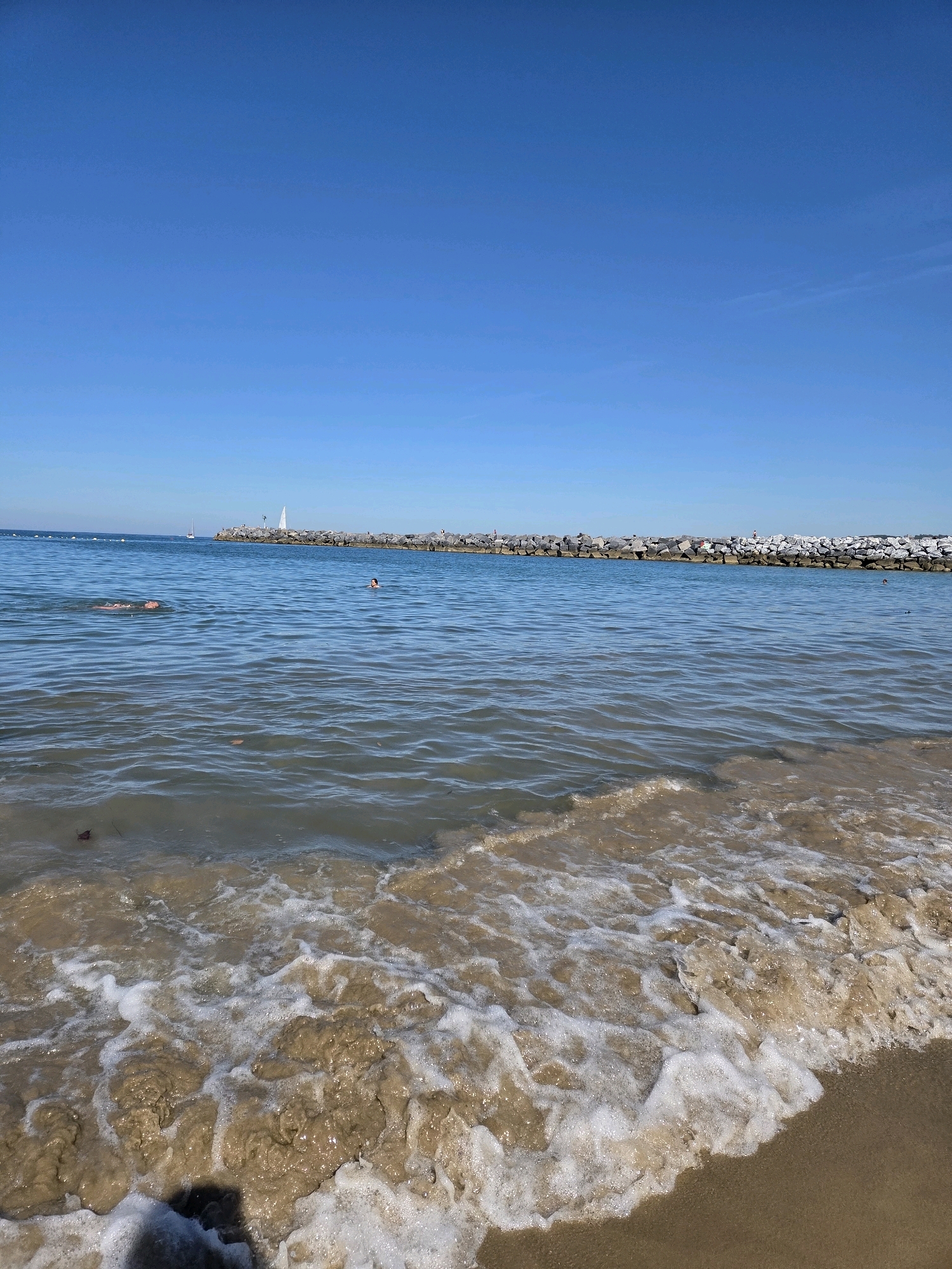 An outdoor, eye-level, wide shot shows a view of the ocean on a sunny day. The ocean water is a clear blue and somewhat choppy, with small waves breaking on the sandy shore in the foreground. In the distance, a rocky breakwater stretches across the horizon, with a few people visible on top of it. A small white sailboat is seen beyond the breakwater. Two individuals are swimming in the ocean; one is further out and appears to be doing the backstroke, while the other is closer to the breakwater and is partially submerged. The sky is a clear, bright blue, indicating good weather. The scene appears to be a public beach or a bay area.