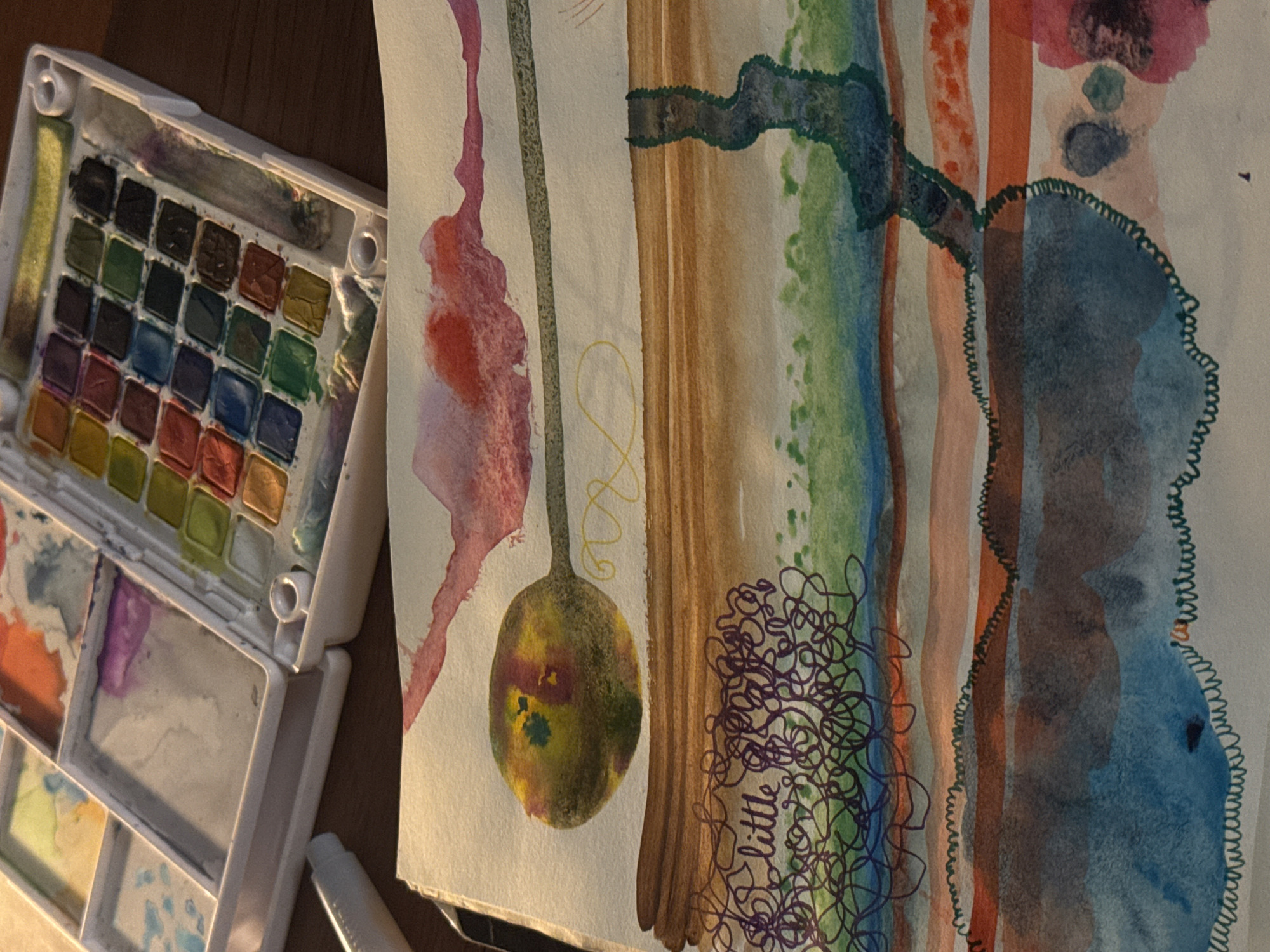 The image shows an overhead view of a watercolor painting and a palette of watercolors. The painting is abstract with horizontal bands of color and some linear elements. There is a prominent circular shape with yellow, green, and red hues in the upper left quadrant. Below this, there are bands of brown, green, blue, and orange. A dark, irregularly shaped vertical element is present in the upper right quadrant of the painting. Some scribbled purple lines and the word "little" are visible on the lower left side. The watercolor palette is a white plastic case with multiple wells containing various colors of dried paint. Some of the wells are wet with pigment. A white tube, possibly of watercolor medium, is also visible to the left of the palette. The objects are placed on a wooden surface, likely a table. The lighting suggests an indoor setting, with a warm glow illuminating the scene. The location is not specified in the image.