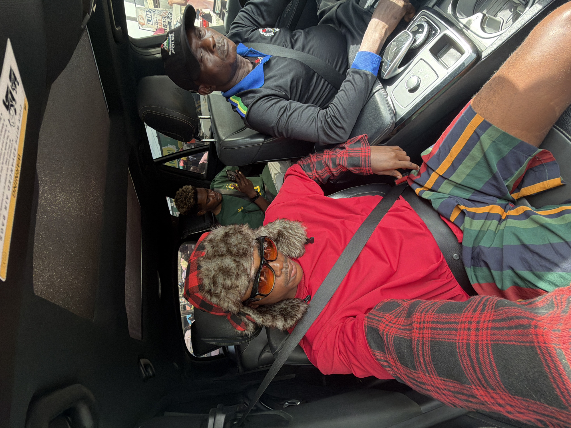 The image captures an interior view of a car during the daytime, showing three men. Bright natural light illuminates the scene, suggesting sunny weather.
In the front passenger seat, a man with dark skin wears a black long-sleeved polo shirt with a blue collar and cuffs. He has a black baseball cap with "POLICE" visibly embroidered on it, and appears to be wearing a seatbelt. His expression is neutral as he looks forward or slightly downwards.
In the back seat, on the right side of the frame, another man with dark skin is seen. He wears a bright red long-sleeved shirt and striped shorts featuring green, blue, yellow, and red patterns. His head is covered by a unique red and black plaid hat with fluffy brown fur ear flaps, and he is wearing orange-tinted sunglasses. A seatbelt crosses his chest, and he appears to be looking towards the left.
Further into the back seat, on the left side, a third man with dark, curly hair is visible. He wears a green shirt and is looking down, seemingly at a smartphone held in his hands.
The car's interior features black leather seats and visible dashboard elements, including a gear shifter and other controls. Through the windows, an urban environment can be glimpsed, featuring buildings and what appear to be posters, consistent with a city setting like Lagos, Nigeria. A warning label on the car's interior, partially visible on the left, reads "WARNING ADVANCED AIR BAGS". The overall atmosphere suggests the men are traveling or resting inside the vehicle.