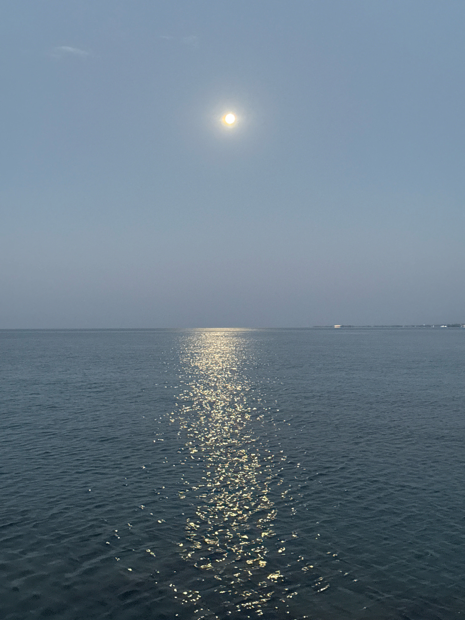 The image depicts a serene, wide-angle view of a tranquil body of water, likely a sea or large lake, under a hazy, pale blue sky. A bright, luminous sphere, possibly the sun or moon, is positioned high in the sky, slightly off-center to the left. Its light casts a shimmering, golden path across the water's surface, creating a reflective trail that stretches towards the viewer. The water itself is a deep, muted blue, with subtle ripples indicating a gentle movement. The horizon line is distinctly visible, separating the water from the sky, and in the far distance, faint shapes of land or structures can be discerned, suggesting a coastal or island setting. The overall atmosphere is one of peacefulness and natural beauty, with the play of light on the water being a prominent focal point. No individuals or specific urban elements are clearly identifiable, giving the scene a timeless and universal quality. The city of Bosteri, Kyrgyzstan, is not depicted in this image; the scene is clearly coastal, not characteristic of Kyrgyzstan's geography.