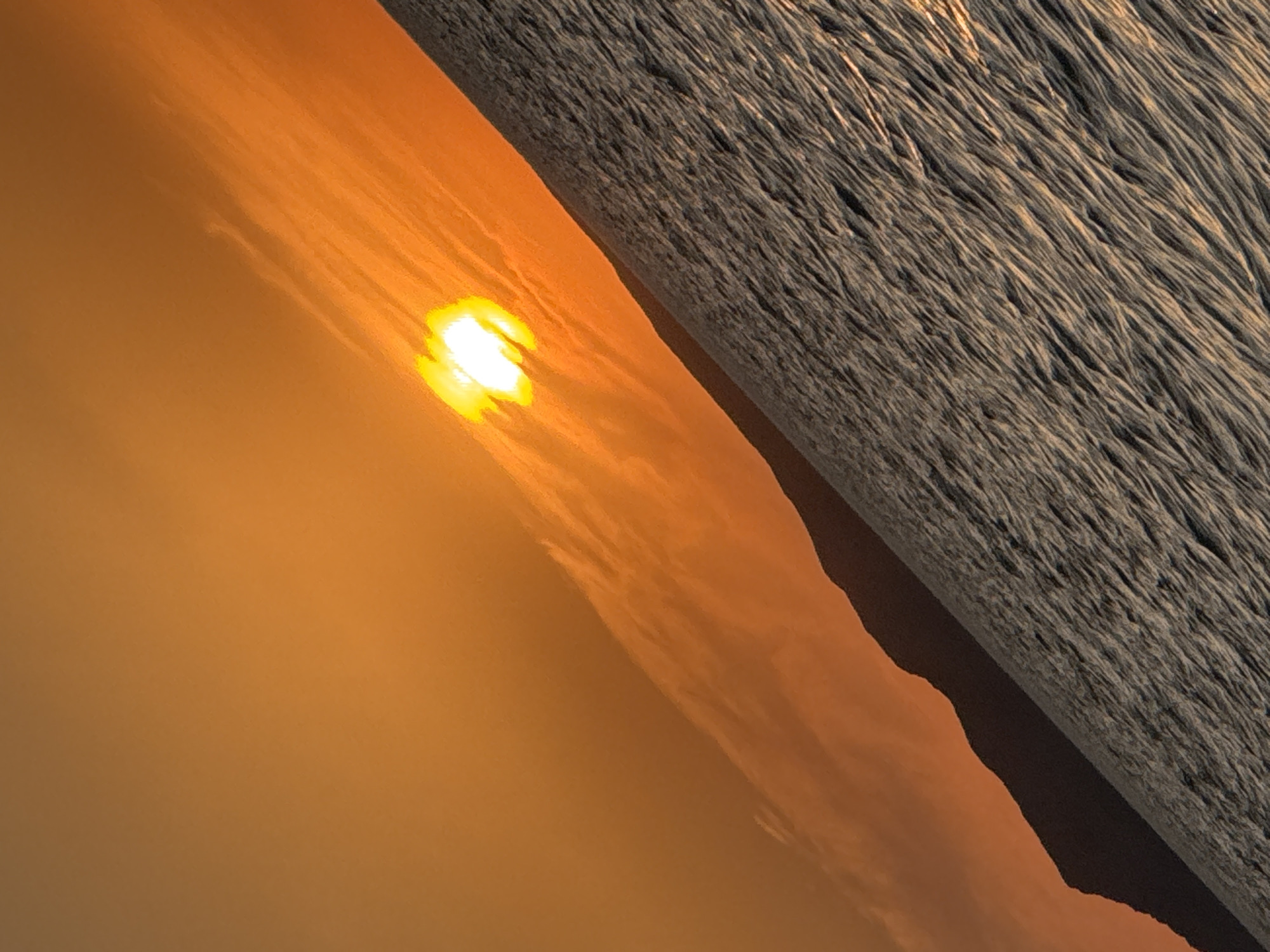 A sunset over the water is depicted in this vertical, angled photograph. The sun, a bright, glowing orb, is positioned in the upper left quadrant of the frame, casting a warm, orange hue across the sky and some wispy clouds. The foreground is dominated by rippling water, appearing textured and reflecting the sunset's light in places. A dark, silhouetted mountain range runs diagonally across the lower portion of the image, separating the water from the sky. The scene is tranquil and natural, with no visible people or man-made structures. The location is specified as Zharbulak, Kazakhstan.