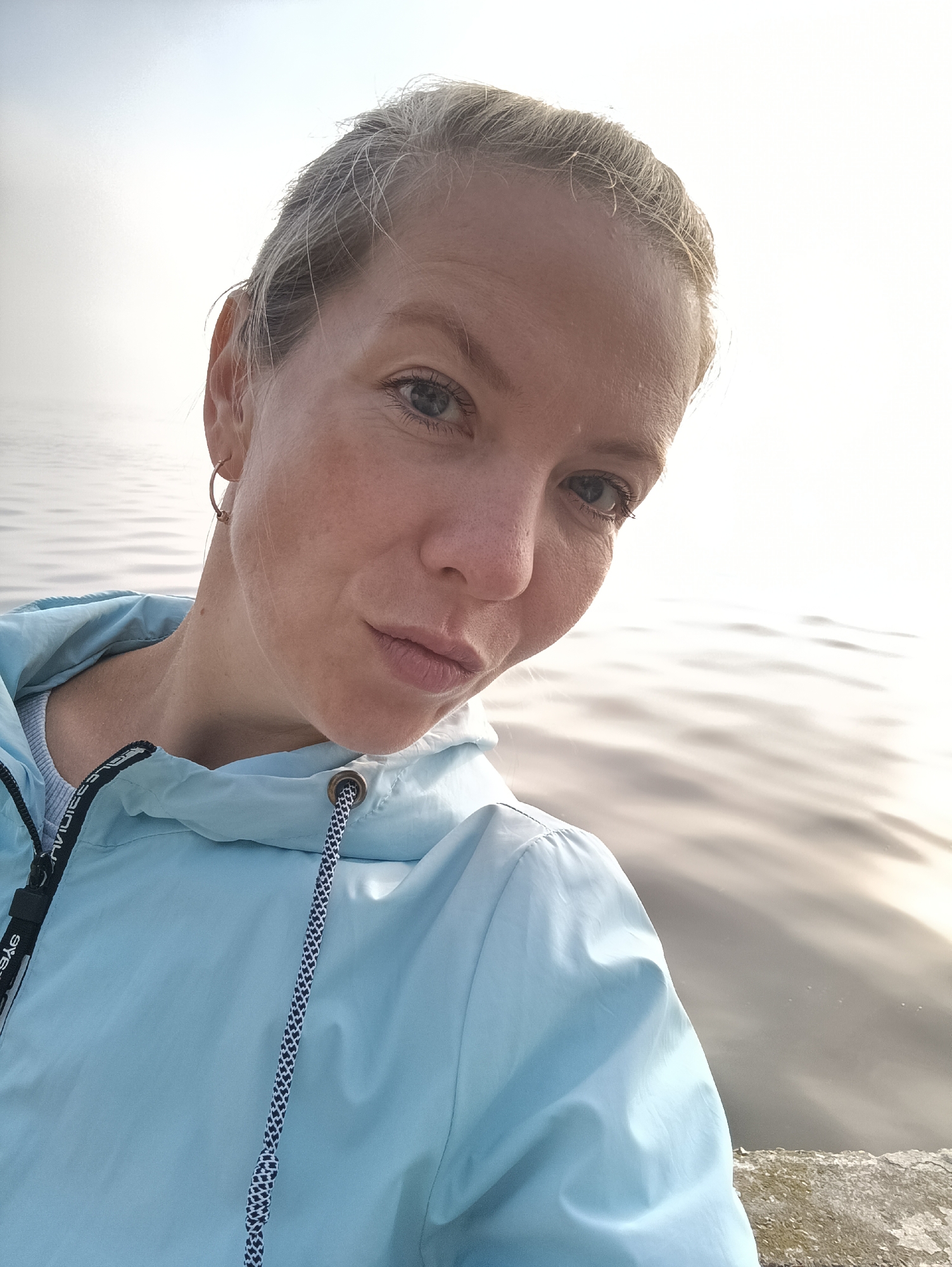 A close-up, selfie-style portrait shows a woman with blonde hair pulled back, facing forward and slightly to the right. She is wearing a light blue jacket with a drawstring hood and has a hoop earring in her visible ear. The background is dominated by the calm, rippled surface of a body of water, possibly a river, and a hazy sky. The lighting suggests it is either early morning or late evening. The woman's expression is neutral, with her lips slightly parted. The image appears to have been taken outdoors, near the water's edge, with a rough, concrete-like surface visible in the lower right corner.