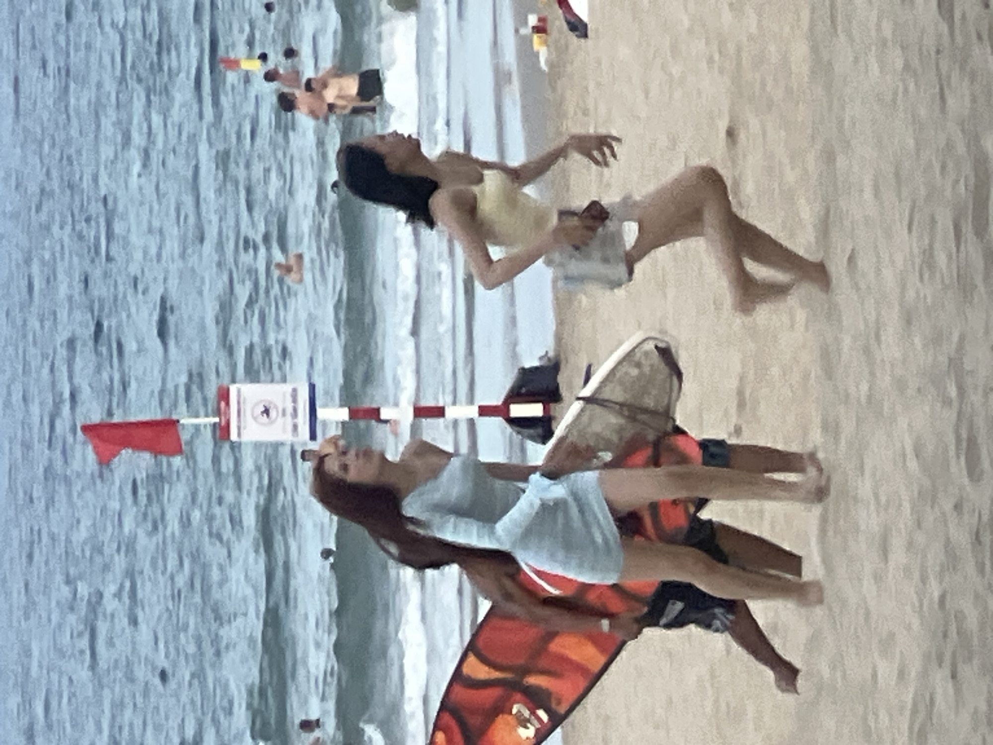 On a sandy beach in Da Nang, Vietnam, two women are depicted near the waterline. The woman on the left, with long dark hair, wears a light blue, one-shoulder dress and is holding a surfboard with an orange and red abstract pattern. She is positioned in front of a partially obscured individual. To the right, a woman with dark hair, wearing a yellow tank top and gray shorts, is captured mid-stride, her right leg extended. In the background, the ocean is visible with waves and several people swimming and standing in the water. A red flag flies from a pole on the beach, next to a white sign with a circular emblem and text. The overall scene suggests a casual day at the beach.