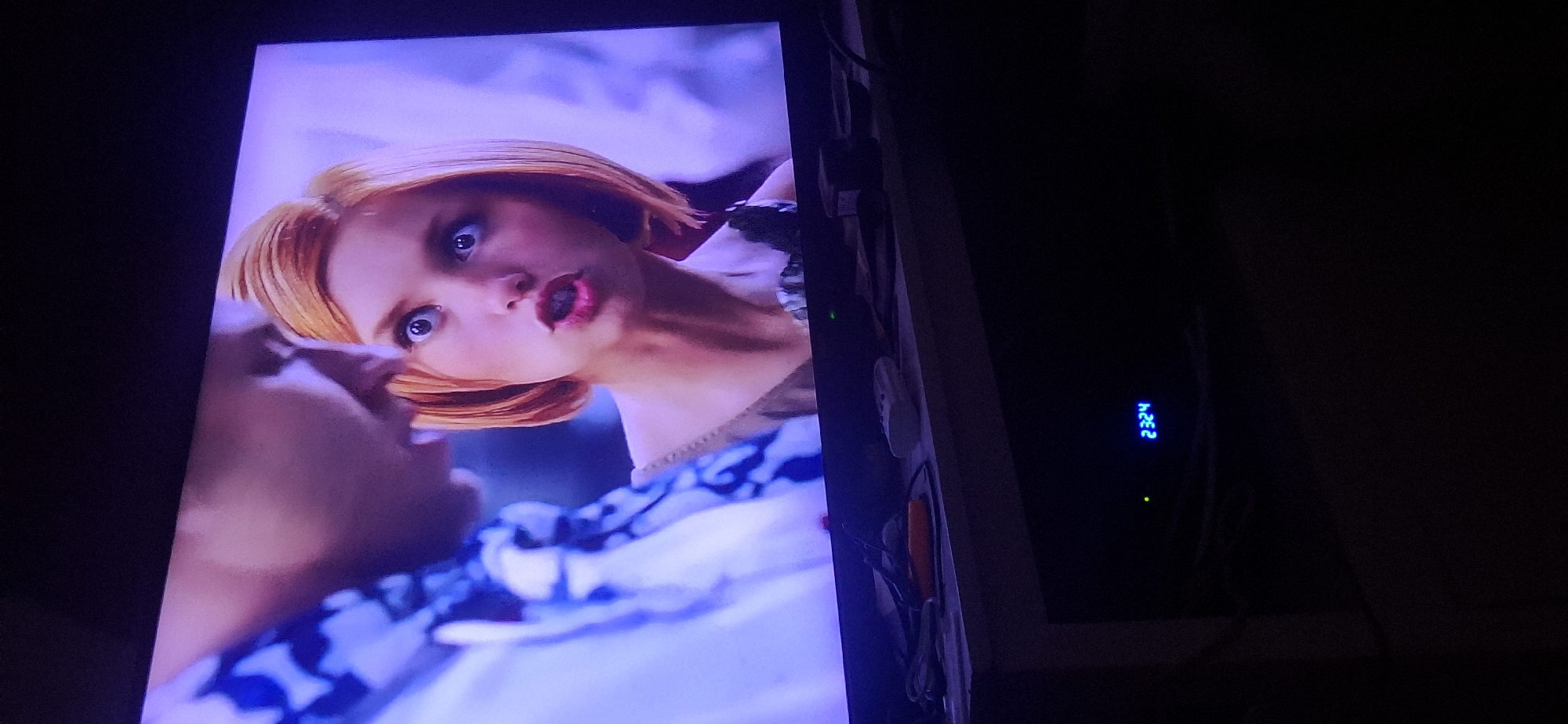 A television screen displays a close-up of a woman with short, blonde hair and wide, blue eyes. Her mouth is open as if speaking or surprised. She is looking towards the left side of the frame, where a portion of another person's shoulder and head is visible. The background on the screen is blurred and appears to be a domestic interior. Below the television, on a dark surface, a digital display shows "2324". The environment suggests a home entertainment setup, with cables and a remote control visible on a shelf in front of the television. The image does not contain any information about the city of Yerocham, Israel.