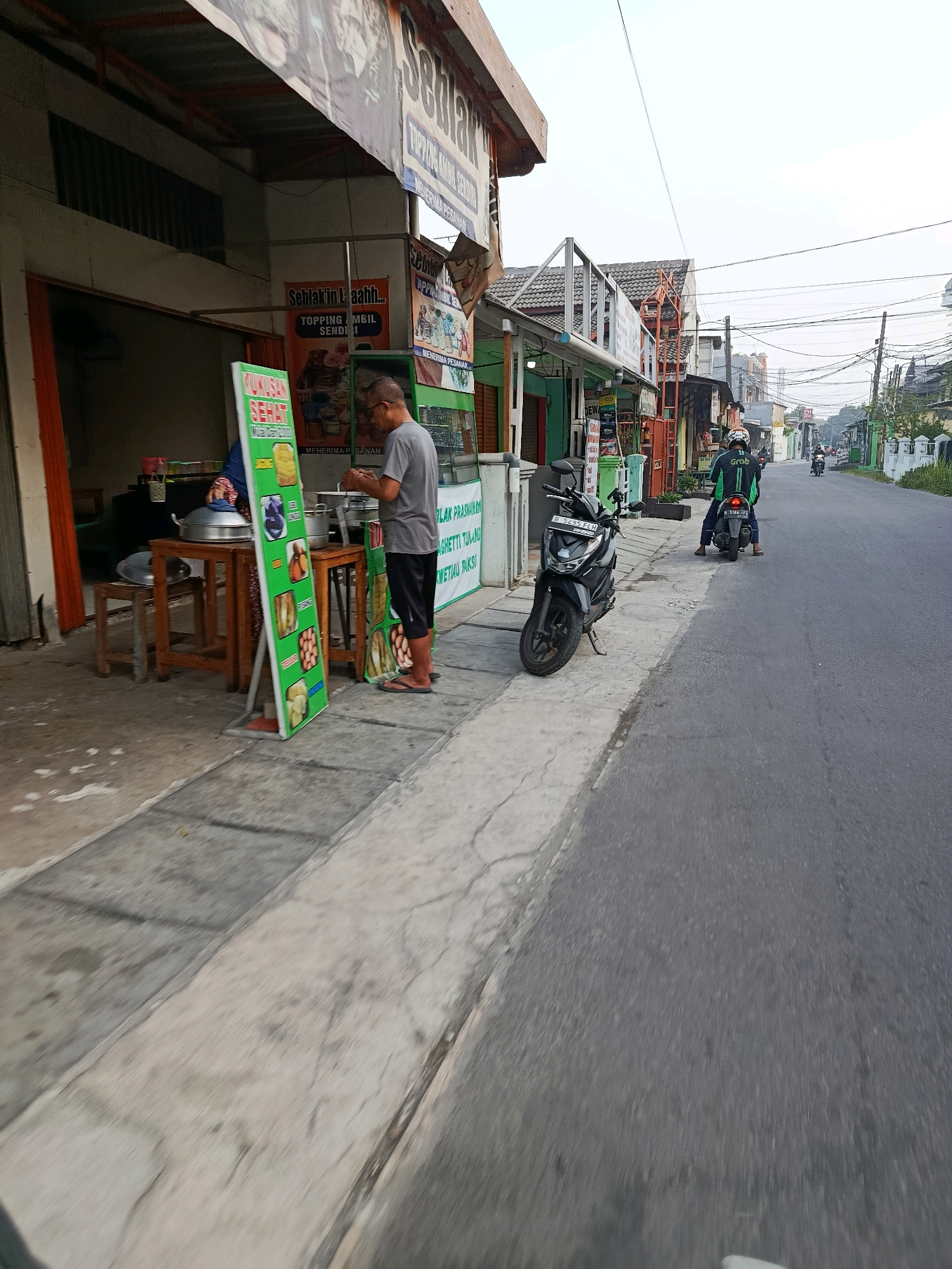The image captures a street scene in Bekasi, Indonesia, likely near a local eatery. In the foreground, a man in a grey t-shirt and black shorts is standing by a food stall. He appears to be interacting with the food preparation or serving area. A motorcycle is parked on the sidewalk to the left of the man, a common mode of transport in the region.
The food stall itself is open-air, with a green menu board displaying various food items and promotions, including "SEBLAK" and "TOPPING AMBIL SENDIRI" (seblak with self-service toppings). Several cooking vessels and a table are visible. To the right of the food stall, the street continues, lined with more small businesses and houses. Further down the street, two people on a motorcycle, one wearing a green jacket with "Grab" branding, are riding away from the camera. The sky is overcast, suggesting it might be early morning or late afternoon. The overall impression is of a typical, bustling neighborhood commercial area in Bekasi.