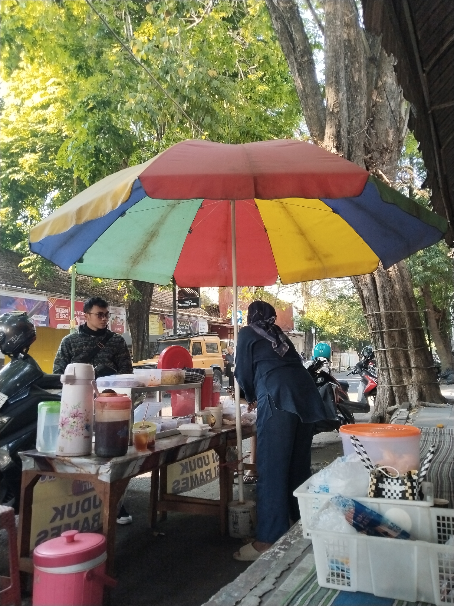 A street vendor stall is set up beneath a colorful, segmented umbrella. A man wearing glasses and a patterned jacket stands behind a table with various containers, including thermoses and a container of iced tea. He is looking to his right. A woman in a dark blue headscarf and matching outfit is standing on the other side of the stall, facing away from the camera and towards the table, where she appears to be attending to something. Several motorcycles are parked nearby. In the background, there is a yellow vehicle, trees with thick trunks and green foliage, and buildings with signage, including a sign for "TOKO SUMBER URIP". The stall is on a paved surface, likely a street or sidewalk, with a curb visible. A large pink insulated container is placed on the ground in the foreground to the left, and two white plastic baskets are in the foreground to the right, one containing items wrapped in plastic. The scene is illuminated by natural sunlight.