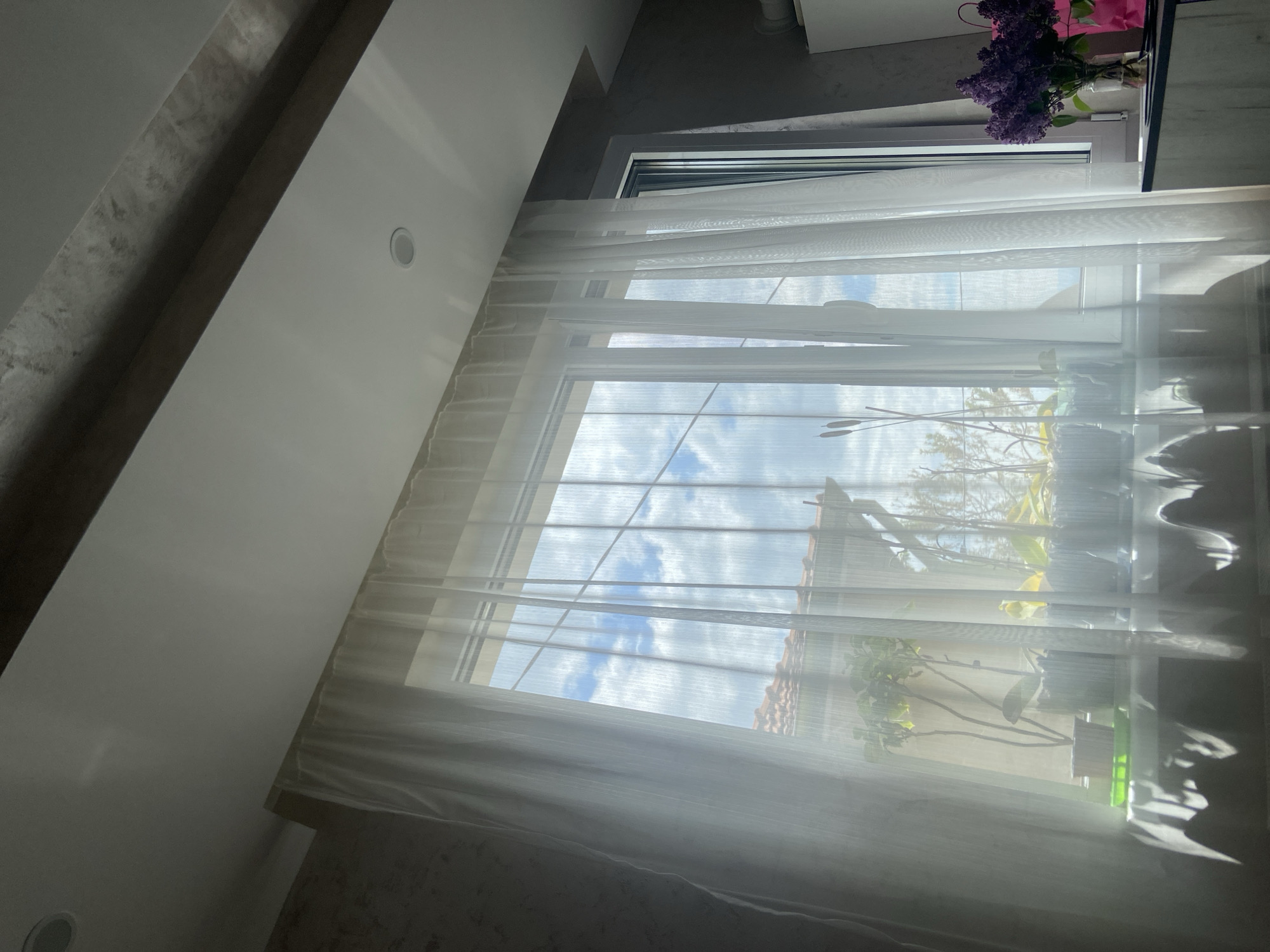 A slightly tilted interior shot captures a window with white sheer curtains, allowing a view of the outside. Through the window, a blue sky with white clouds can be seen along with a building with an orange tile roof and green foliage suggesting a garden or trees. On the windowsill, various potted plants are visible, and a vase of purple flowers is on the upper right corner, near a dark object that is slightly out of focus. The interior reveals a section of a white wall and ceiling, angled due to the tilted perspective. The overall lighting suggests a bright, sunny day. No people are present in the image. There is no evidence to prove it is located in Dragovishtitsa, Bulgaria.
