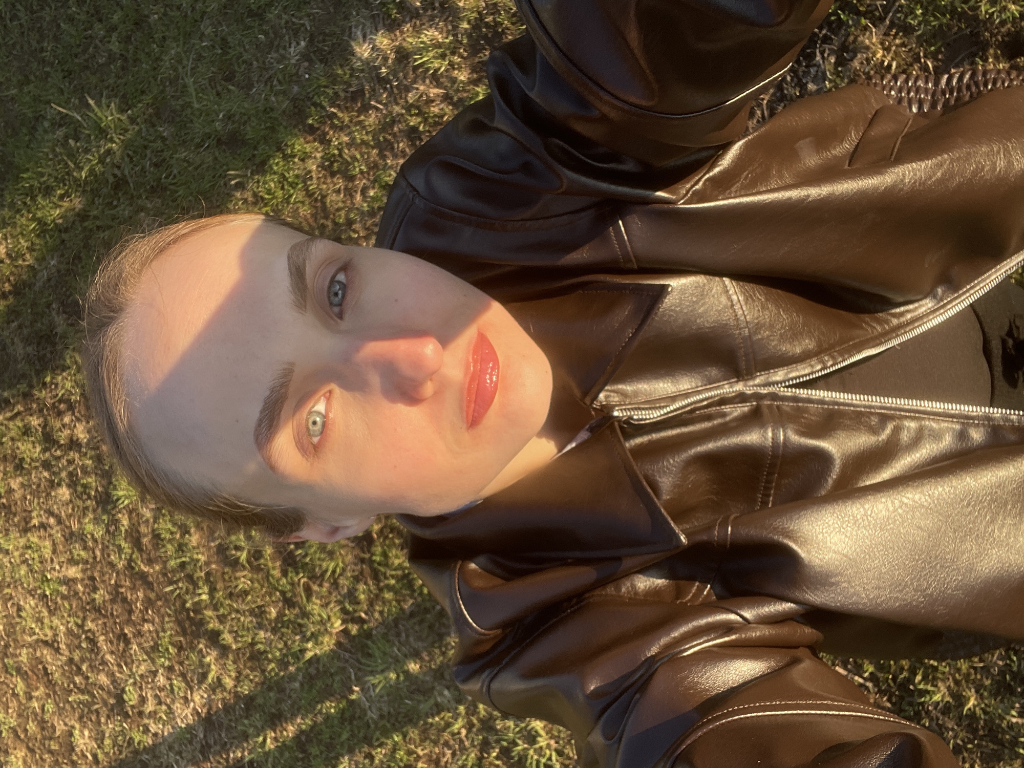 A tilted, high-angle portrait captures a young individual looking directly at the camera. The subject is positioned diagonally across the frame, with their head in the upper left and their body extending towards the lower right. They are wearing a shiny, dark brown leather jacket with visible zipper details.
The scene is outdoors, with the background consisting of green and brown grassy ground, suggesting a natural or park-like environment. Strong, warm sunlight, indicative of golden hour or late afternoon, illuminates the right side of the subject's face, creating a bright highlight across their forehead, nose, and cheek. The other side of their face is in shadow. Their eyes are a striking blue or grey, and their lips are adorned with glossy, reddish-pink lipstick. The expression is neutral, with a direct gaze. No specific activity beyond posing for a photo is depicted.
The location context indicates this photo was taken in Sovetsko-Nikol'skoe Lesnichestvo, Russia, which aligns with the natural outdoor setting suggested by the grass. There is no visible text in the image.
