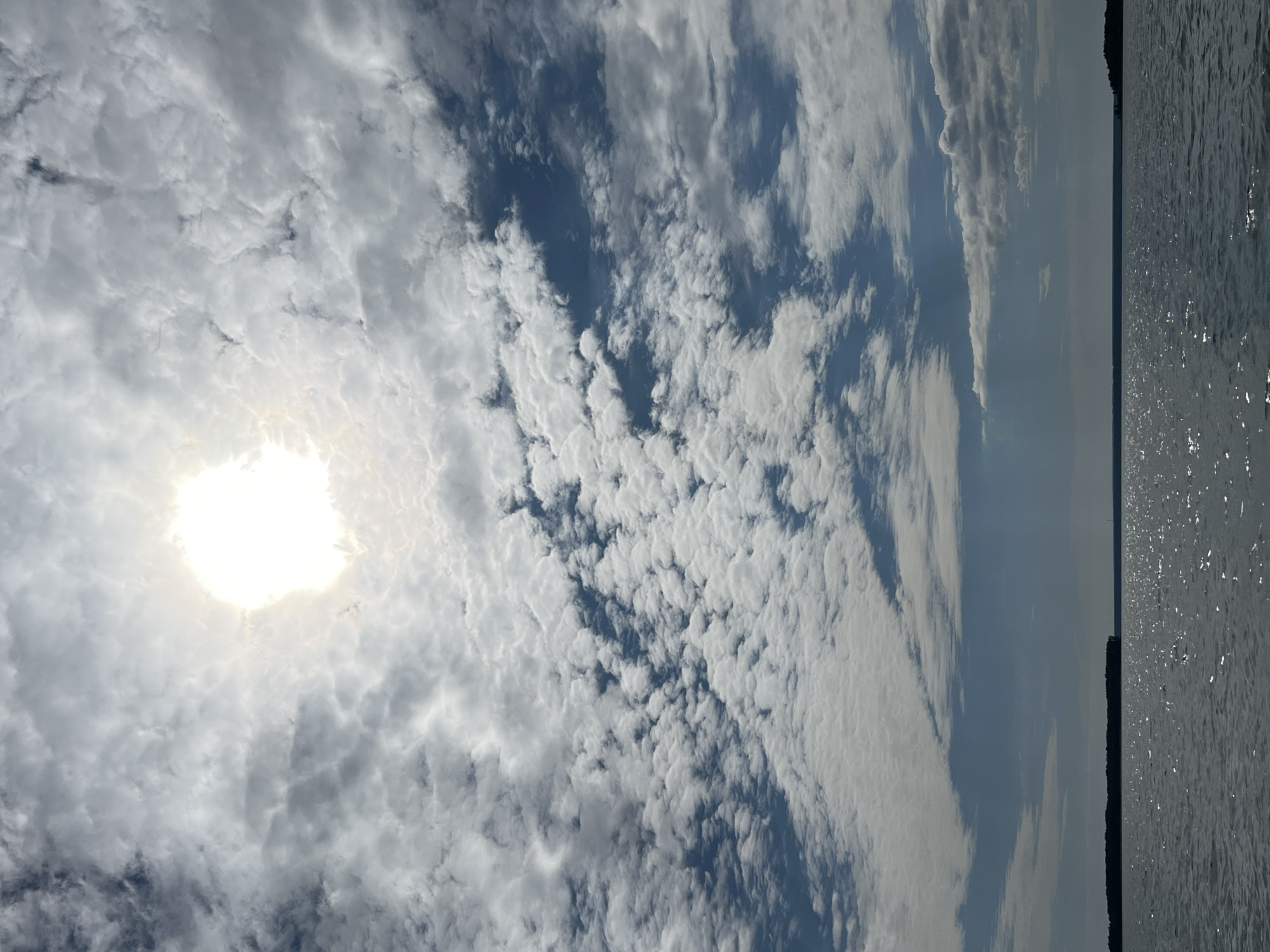 The image depicts a vast expanse of water under a cloudy sky. The sun is partially obscured by clouds, creating a bright halo effect. The water appears calm with a gentle ripple, reflecting the light from the sun. In the distance, a dark, silhouetted landmass runs horizontally across the frame, suggesting a shoreline or an island. The overall atmosphere is serene and natural. There are no people, animals, or man-made structures visible in the image, apart from the implied landmass in the distance. The location, Novopichugovo, Russia, is a rural settlement on the banks of the Yenisei River, known for its natural beauty and vast landscapes. This image captures the essence of such a location, showcasing the interplay of water, sky, and land.