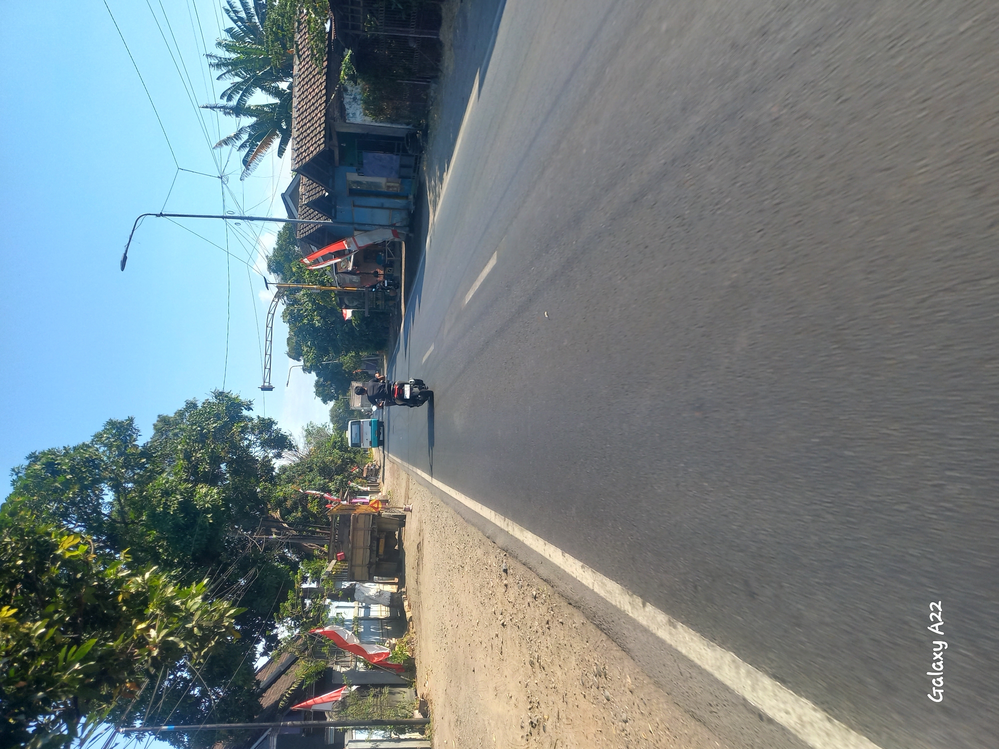 A daytime exterior scene shows a paved road in Kotakan, Indonesia. A person rides a dark-colored motorcycle away from the camera in the right lane. To the left of the road, there is a dirt shoulder. Behind the dirt shoulder are several Indonesian flags displayed on poles, a truck with a wooden bed, and some buildings. A light-colored bus travels in the opposite direction on the road. In the background, there are trees and palm trees, and a bright blue sky. The watermark "Galaxy A22" is visible in the bottom left corner of the image.