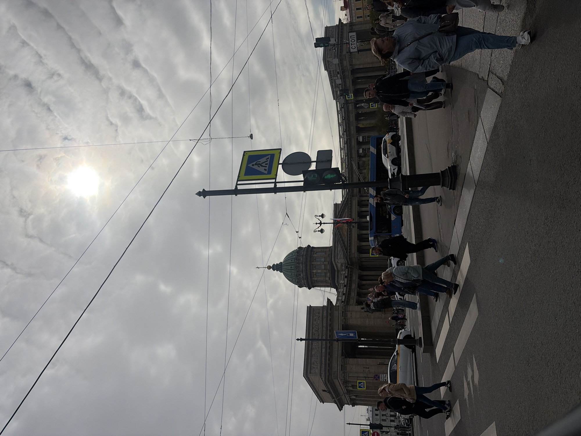 The image shows a street scene in Saint Petersburg, Russia, featuring the Kazan Cathedral in the background. Pedestrians are crossing the street at a zebra crossing with a crosswalk sign. A traffic light is visible and it is green. The sky is overcast with clouds and the sun is visible in the top left of the image. Overhead wires run across the scene. Several buildings can be seen including the Kazan Cathedral.