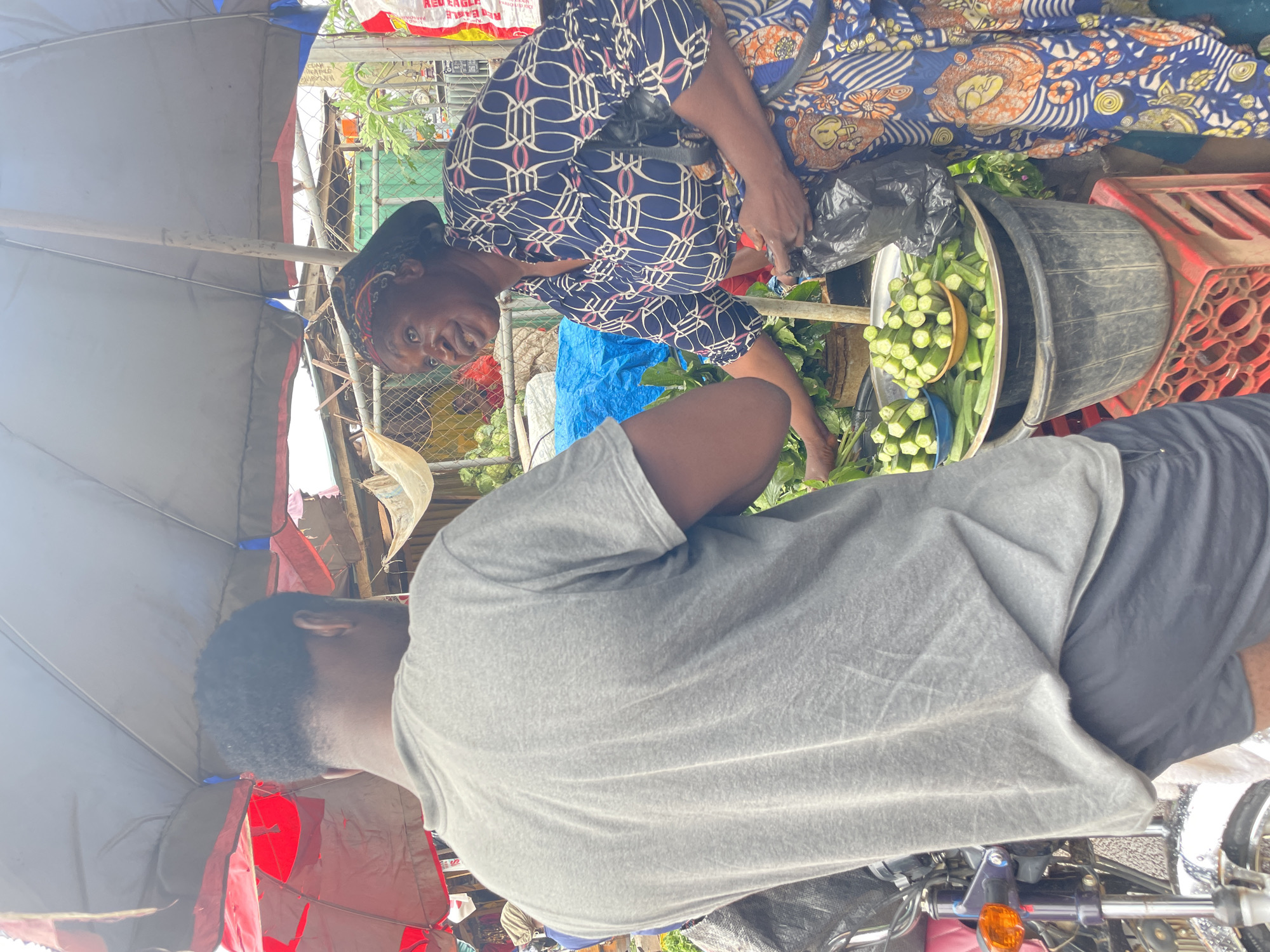 In Akwanga, Nigeria, two individuals are engaged in a transaction at a market stall. A woman, wearing a blue and white patterned headscarf and a blue and white patterned dress with an orange and brown patterned wrap skirt, stands behind a display of fresh produce. She holds a black plastic bag in her right hand and is gesturing with her left hand towards a bowl of sliced okra and whole okra pods. In front of her, a man, wearing a grey t-shirt and dark shorts, faces away from the camera, his back to the viewer, with his left hand extended as if receiving or selecting the produce. The stall is partially covered by a large, faded grey umbrella with red trim. Beneath the produce, a large black plastic tub sits on a red plastic crate. A motorcycle is partially visible to the left of the frame, with its handlebars and orange turn signal illuminated. In the background, a chain-link fence and various market structures are visible, suggesting a bustling urban market environment.