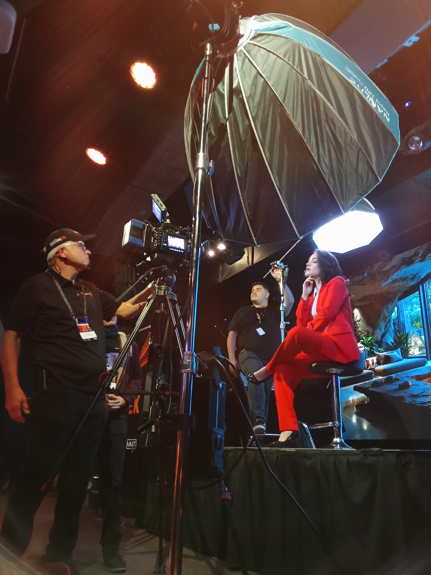 The image captures a behind-the-scenes view of a professional video recording setup. In the foreground, a man wearing a black polo shirt and a baseball cap operates a professional video camera mounted on a tripod. He appears focused on adjusting the camera. To his right, a woman in a vibrant red suit is seated on a stool, looking off to the side with her hand resting near her chin, suggesting she is being filmed or is awaiting her cue.
A large softbox light, angled to illuminate the woman, is a prominent feature in the scene. In the background, two other individuals are visible, one standing behind the seated woman and another further back, both appearing to be part of the production crew. The environment seems to be an indoor studio or event space, indicated by the lighting equipment and the artificial rock formations and greenery visible in the background, which suggest a themed set. The overall impression is one of a professional media production in progress.