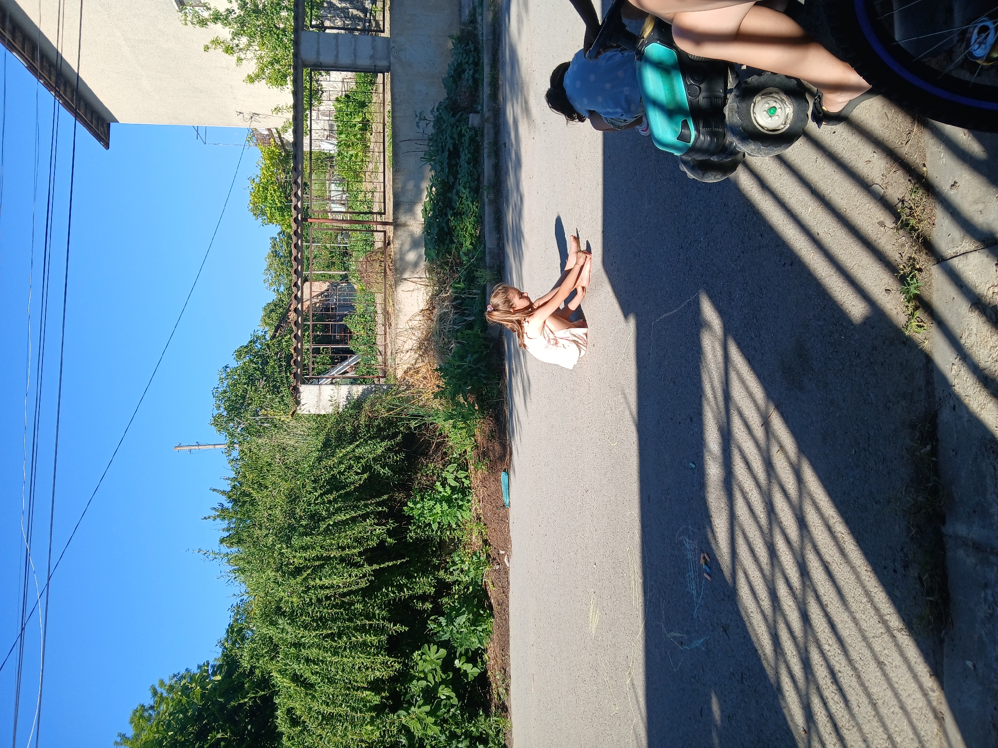 A young girl with blonde hair pulled into a ponytail, wearing a light-colored dress, sits on the asphalt street in front of a dense green hedge and a metal fence. She is facing left, with her legs extended and her hands reaching towards the ground. Behind her and to the right, another child wearing a blue and white polka-dot shirt is crouched down next to a blue plastic toy tractor. The sun casts long, diagonal shadows across the street, indicating it is likely late afternoon. Chalk drawings, including a circle and some colorful lines, are visible on the asphalt near the children. The background shows a residential area with a building and trees. The location is identified as Obrochishte, Bulgaria.
