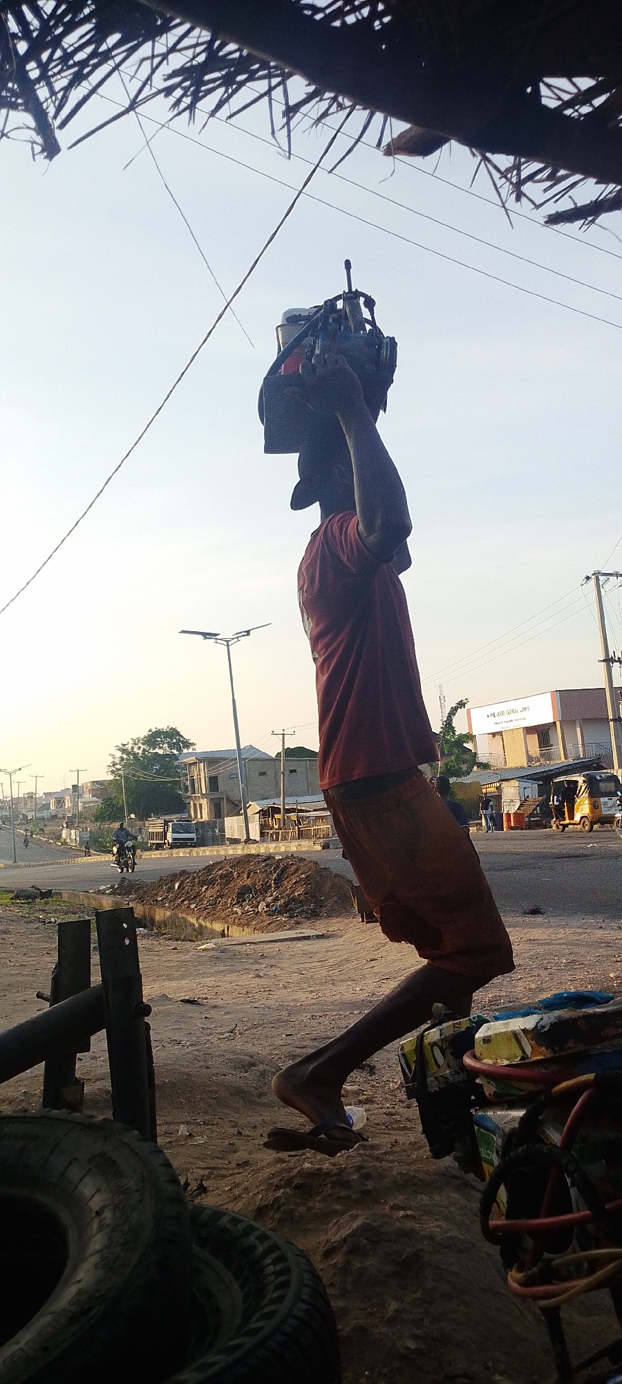 A man in a red shirt and reddish-brown shorts carries a heavy, unidentified object on his head. He is wearing flip-flops. The environment appears to be a roadside location, possibly in Lokoja, Nigeria, as per the prompt. The ground is dusty and uneven. There are tires stacked near him, as well as a piece of equipment with wires. The background shows a road with buildings, streetlights, and vehicles, including a motorcycle and an auto rickshaw. The sky is a pale blue, suggesting dawn or dusk.