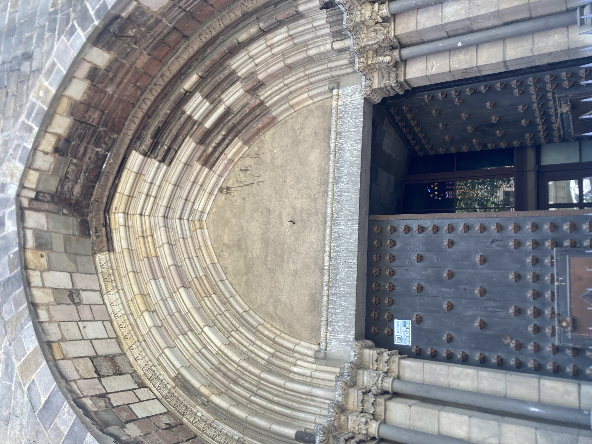 The image shows a close-up view of the entrance to a historic building in Barcelona, Spain. The entrance features a large, arched doorway with ornate stonework. The arch is made up of multiple concentric rings of carved stone, with decorative elements on the inner and outer edges. Above the arch, the facade is constructed with a mix of rough-hewn stones.
The doorway itself is made of heavy, dark metal, studded with numerous decorative metal bosses arranged in a grid pattern. The doors are partially open, revealing a glimpse of stained glass windows inside, reflecting some colorful light. The architectural style suggests a religious building, possibly a cathedral or church, due to its grandeur and the presence of what appears to be religious iconography in the stained glass.
On the left door, a small white sign with a blue border and text is visible. It appears to be an alert or information sign, with the word "ATRAS" prominently displayed along with a phone number. The overall impression is one of age and historical significance, with a sense of weathered grandeur.