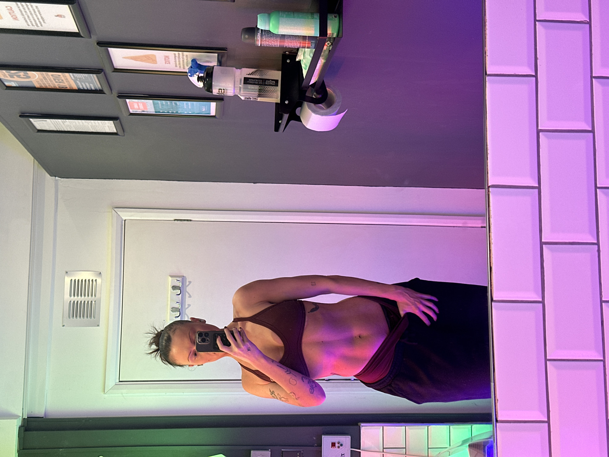 A person with a dark bun is standing in front of a bathroom mirror, holding a phone up to take a picture of their reflection. The person is wearing a brown sports bra and dark sweatpants, revealing their torso. Tattoos are visible on their arms. The bathroom has grey walls, white subway tiles, and a white door with a vent. A toilet paper holder with bottles and framed pictures are mounted on the wall behind the mirror. The scene is illuminated with colorful lights, with green at the bottom and purple near the top. The location is in a bathroom in Tonbridge and Malling, United Kingdom.