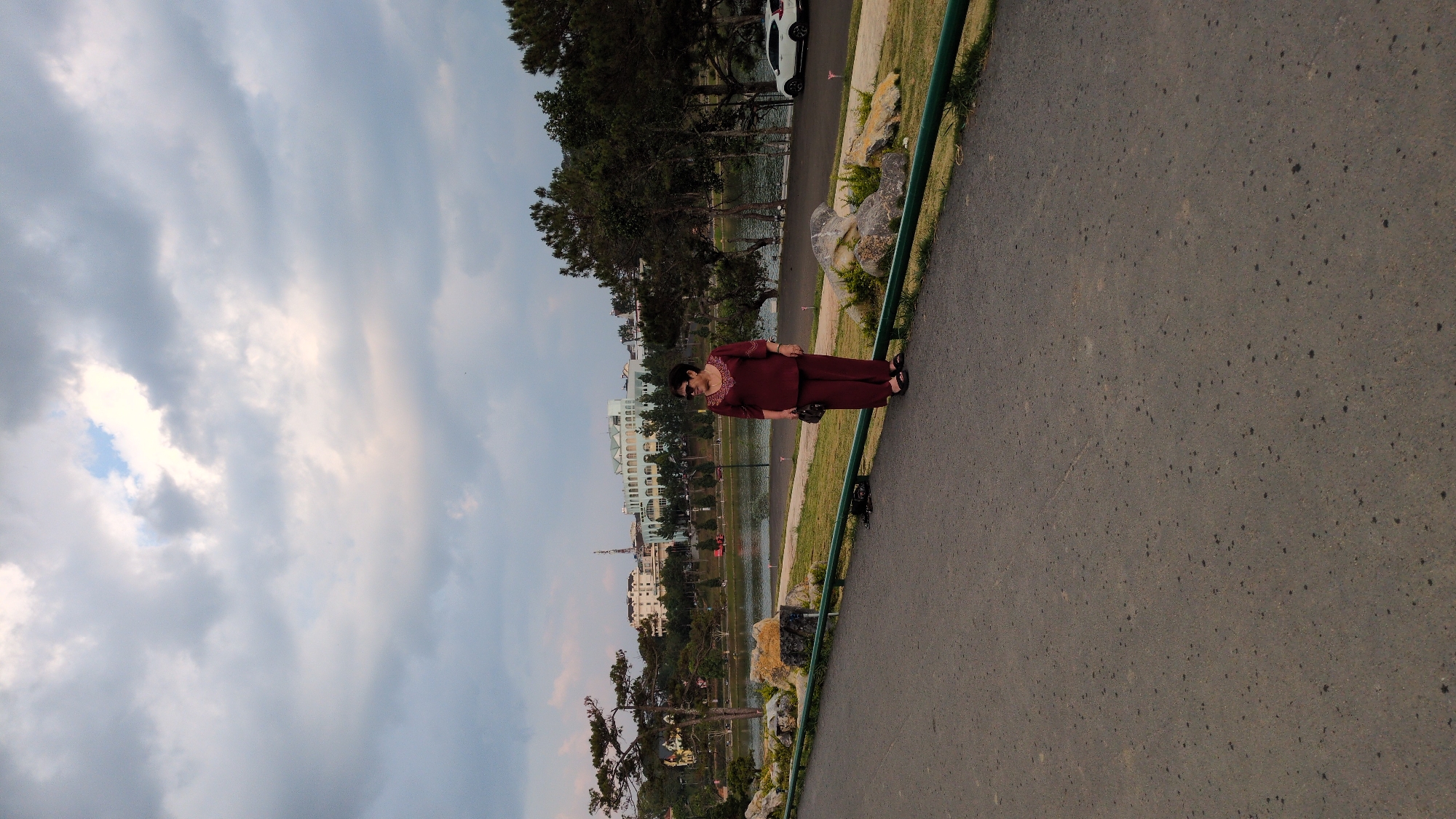 The image depicts a woman standing on a paved path next to a body of water. The woman is wearing a maroon-colored outfit. In the background, there are trees, a white car, a building, and a cloudy sky. The image was taken in Da Lat, Vietnam.