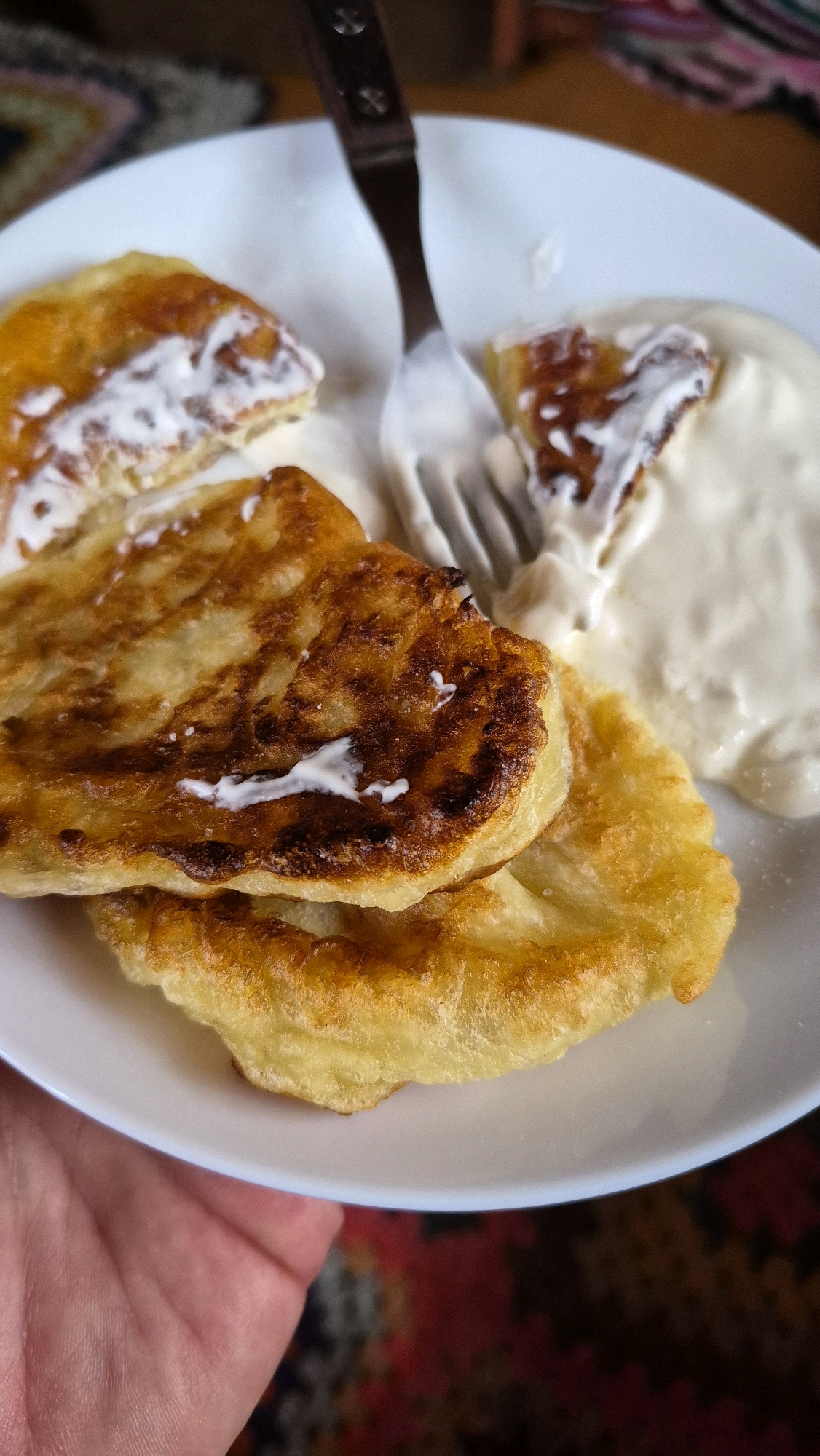 A close-up, top-down view of a white plate holding several golden-brown pancakes, some topped with dollops of white sour cream or yogurt. A fork with a wooden handle is partially submerged in the sour cream, with a bite taken out of one of the pancakes on the right side of the plate. The pancakes appear fluffy and slightly greasy, with some darker, caramelized edges. A hand with visible skin texture is holding the plate from the bottom left of the frame. The background consists of a blurred, colorful textile, possibly a rug or tablecloth. The setting is indoors, likely a dining area.