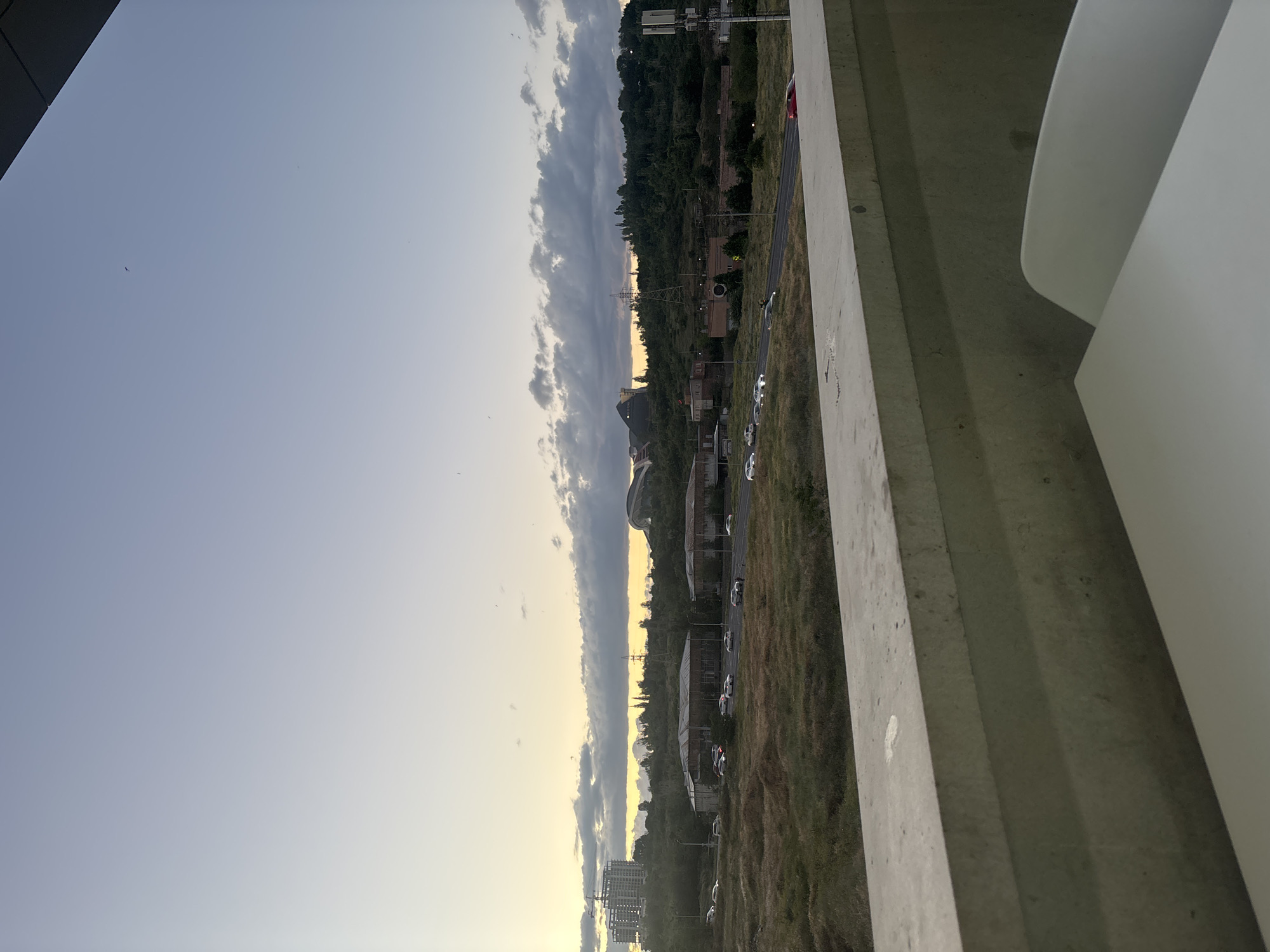This media file presents a panoramic outdoor view of Yerevan, Armenia, likely captured from an elevated position such as a balcony or rooftop. The scene is set during either sunrise or sunset, indicated by the gradient sky transitioning from a soft, pale blue at the top to a warm, yellowish-orange glow near the horizon. A prominent band of dark, elongated clouds stretches horizontally across the middle of the sky.
In the immediate foreground, a substantial concrete ledge or wall occupies the right side, along with a distinct white, curved architectural feature, suggesting the structure from which the photo was taken. Beyond this barrier, a strip of dry, grassy ground leads to a road where several white cars are neatly parked, with a red car partially visible.
The middle ground reveals a blend of urban development and natural landscape. Various buildings, some appearing older or industrial, are interspersed with clusters of trees and vegetation. A construction crane is visible in the far left distance, hinting at ongoing development. Power lines or utility poles punctuate the landscape. Further back, hills covered in lush greenery form a natural backdrop, and a large, possibly dome-shaped or stadium-like structure is discernible on the left horizon. The overall impression is a tranquil, expansive twilight scene, showcasing the city's interface with nature. There are no people visible, no specific activities taking place, and no visible text.