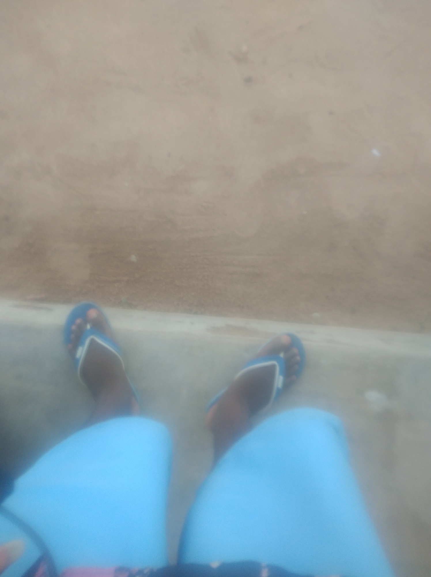 The image shows a low-angle view of a person's legs and feet, with the feet wearing blue flip-flops. The person is wearing light blue pants. The background appears to be a sandy or dusty surface, possibly a dirt road or a beach. The lighting suggests it is daytime. The image quality is low, with significant blur. The specific location in Jalingo, Nigeria cannot be determined from the provided image.