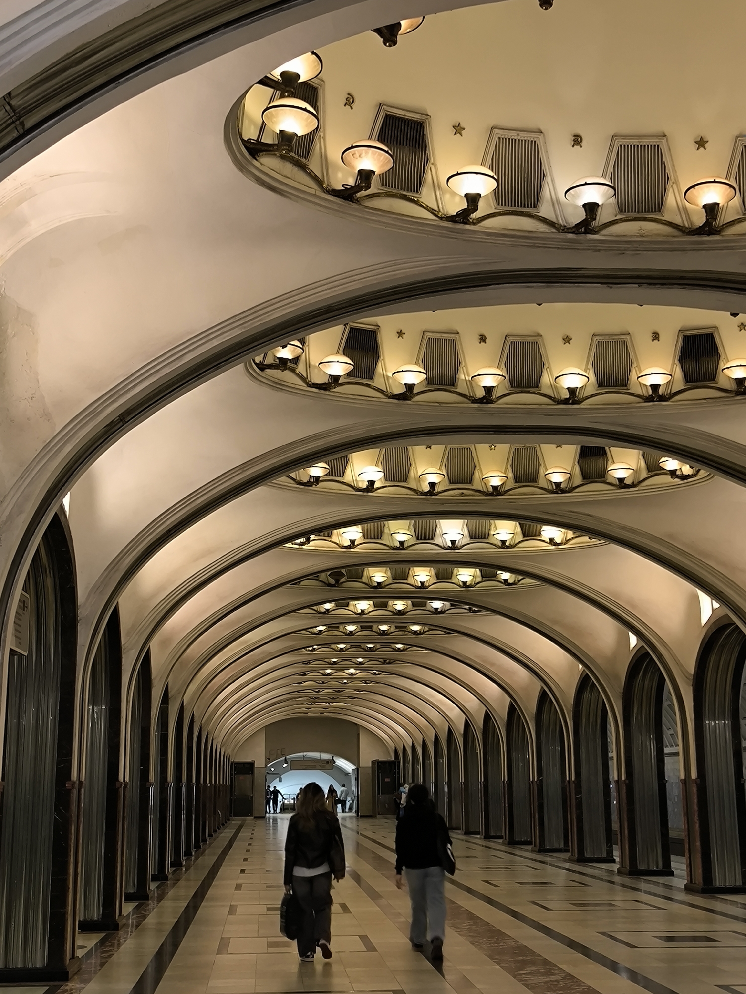 This image depicts an ornate, domed hallway with arched passageways and a series of identical light fixtures on the ceiling. The architecture suggests a grand public space, likely a metro station, given the style and the presence of people walking. The ceiling is adorned with rows of inverted bowl-shaped lamps and ventilation grates, interspersed with small star ornaments. The walls are lined with a repeating pattern of dark, polished columns and recessed arches, creating a sense of depth and grandeur. The floor is tiled in a pattern of light and dark squares with a contrasting stripe down the center.
Two individuals are walking away from the camera down the hallway. The person in the foreground on the right is wearing a dark jacket and light-colored pants, carrying a dark bag. The person in the foreground on the left is wearing a dark jacket and dark pants, also carrying a bag. In the distance, other figures are visible, suggesting this is a busy thoroughfare. The lighting is warm and somewhat dim, typical of an underground environment. The overall impression is one of solemnity and classic architectural beauty.
This scene is characteristic of the Moscow Metro, particularly stations built during the Soviet era, known for their opulent and monumental designs. The specific architectural elements, such as the arched ceilings, decorative lighting, and the use of marble-like finishes, are hallmarks of stations like Mayakovskaya or Ploshchad Revolyutsii. The presence of Soviet-era symbols, like the stars, further points to a Moscow Metro station.