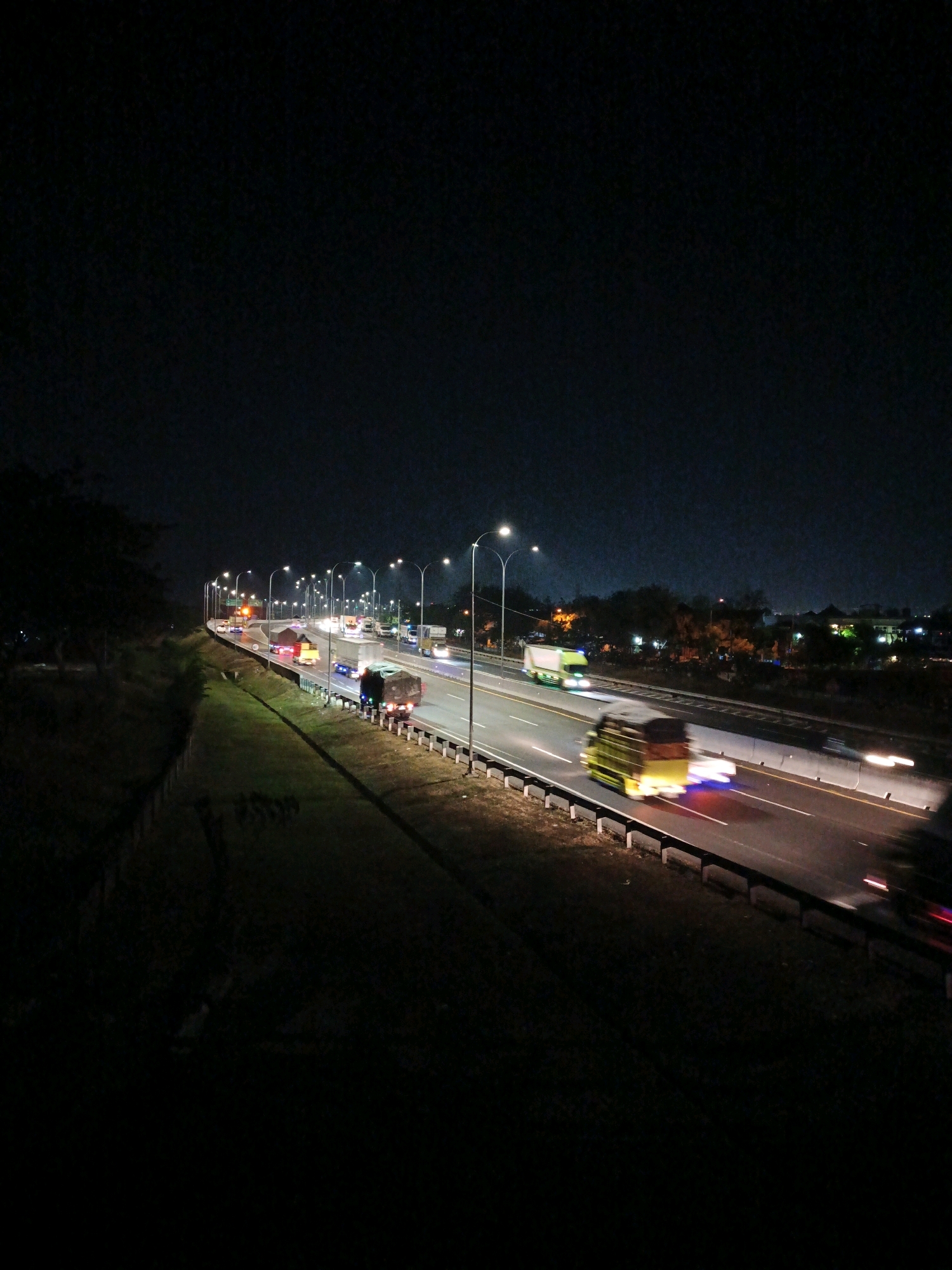 This is a nighttime, long-exposure photograph of a highway in Kludan, Indonesia. The highway is illuminated by streetlights and the headlights and taillights of passing vehicles. Several trucks are visible on the highway, some of which are blurred due to motion. The area adjacent to the highway is grassy, with some trees and distant buildings visible in the background. The sky is dark and clear.
