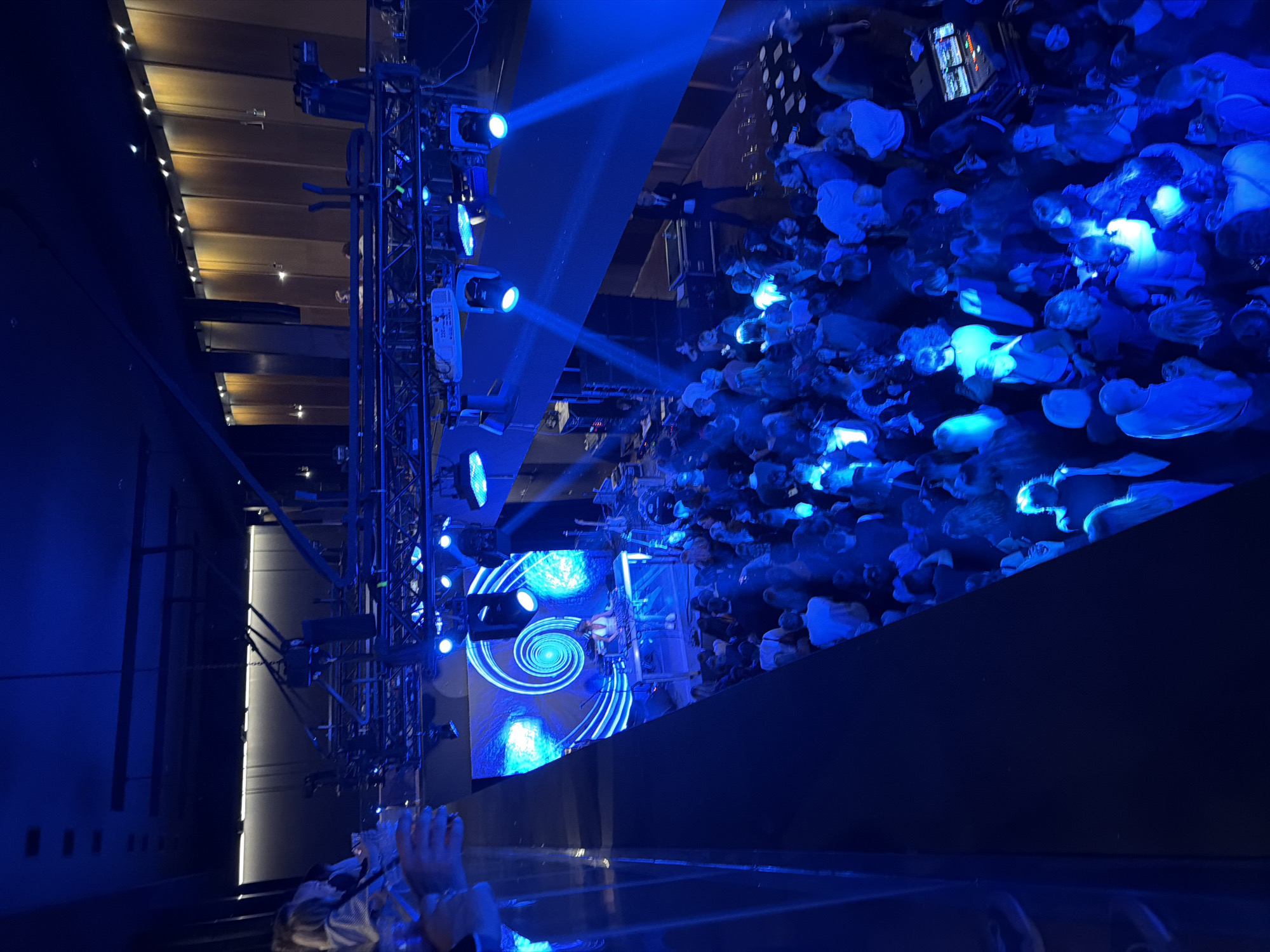 An overhead perspective captures a large-scale indoor event, likely a concert, presentation, or conference, held in a venue in Tbilisi, Georgia. The scene is immersed in vibrant blue lighting, creating a dynamic and engaging atmosphere.
In the mid-ground, a brightly lit stage dominates, featuring a large LED screen displaying a prominent, swirling blue and white spiral graphic. A single individual, possibly a performer or speaker, stands on the stage, facing the audience. To the left of the stage, a robust truss structure supports an array of spotlights and stage equipment, emitting strong beams of blue light that cut through the air.
A vast audience occupies the floor area in front of the stage, densely packed and illuminated by the intense blue lights. Many audience members appear engaged, with some individuals holding up glowing objects or phones, contributing to the overall blue glow. The controlled lighting indicates an evening event or a dedicated indoor performance setting.
Architectural details include a wooden slatted ceiling with embedded lights visible in the upper part of the image, and dark walls framing the event space. The foreground shows a dark barrier, suggesting the photograph was taken from an elevated position, such as a balcony or upper tier, with a few hands visible at the bottom right corner, indicating other observers. No explicit text is visible on screens or signs.